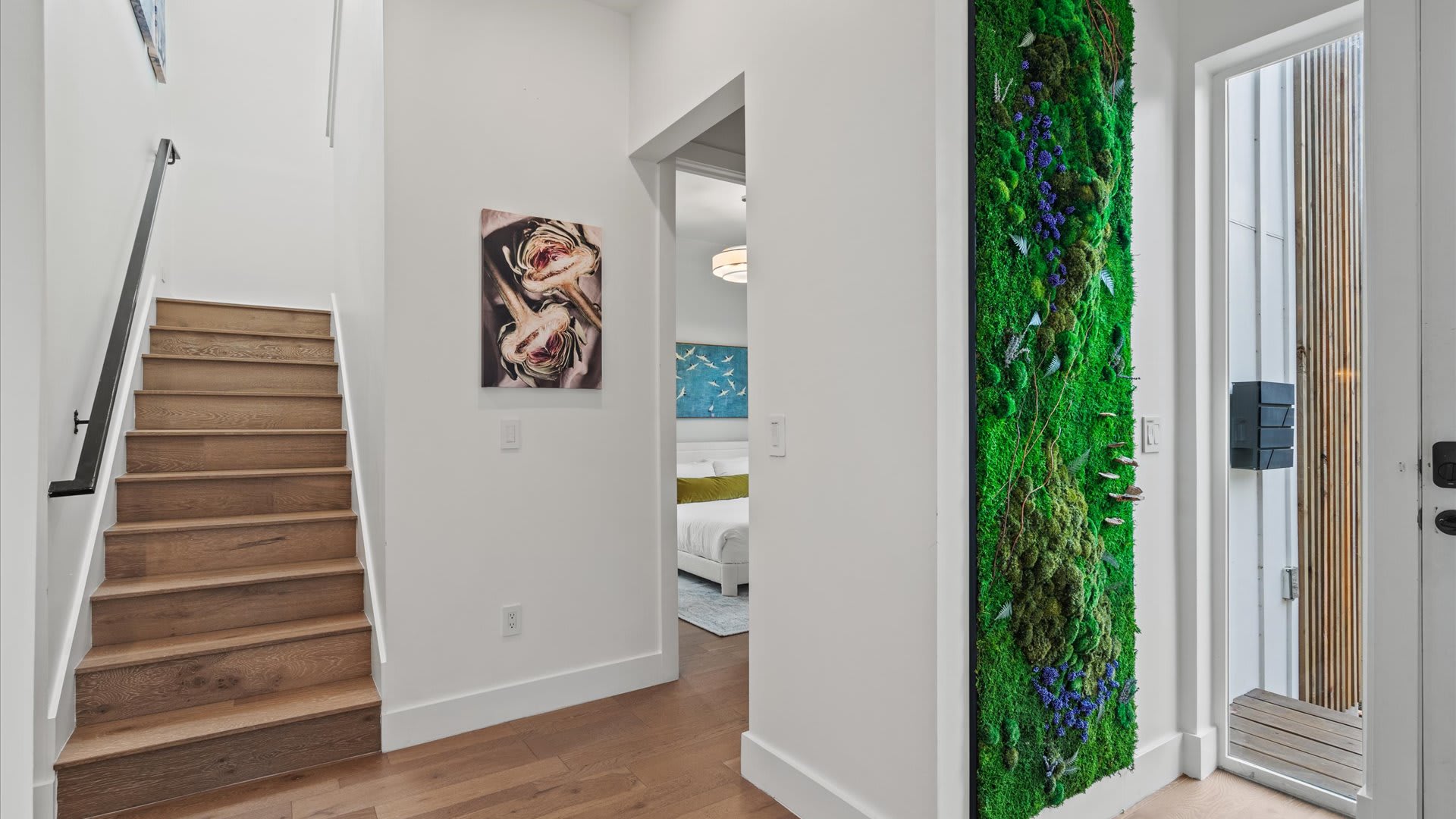 A modern foyer with light hardwood floors, a wooden staircase with black railing, and a striking vertical moss wall art installation next to a doorway leading to a bedroom.