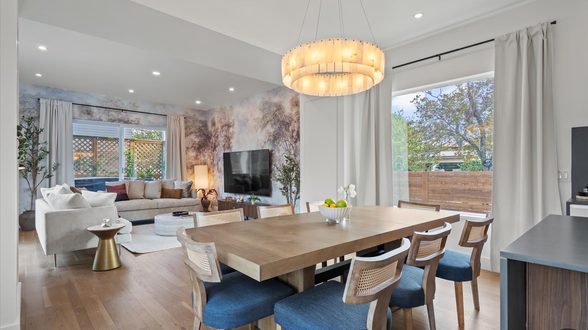Elegant dining area featuring a long wooden table paired with six chairs, each boasting cane backs and blue upholstered cushions. A large, circular chandelier with a textured finish hangs above, illuminating the space that opens into the living room with a decorative mural wall.