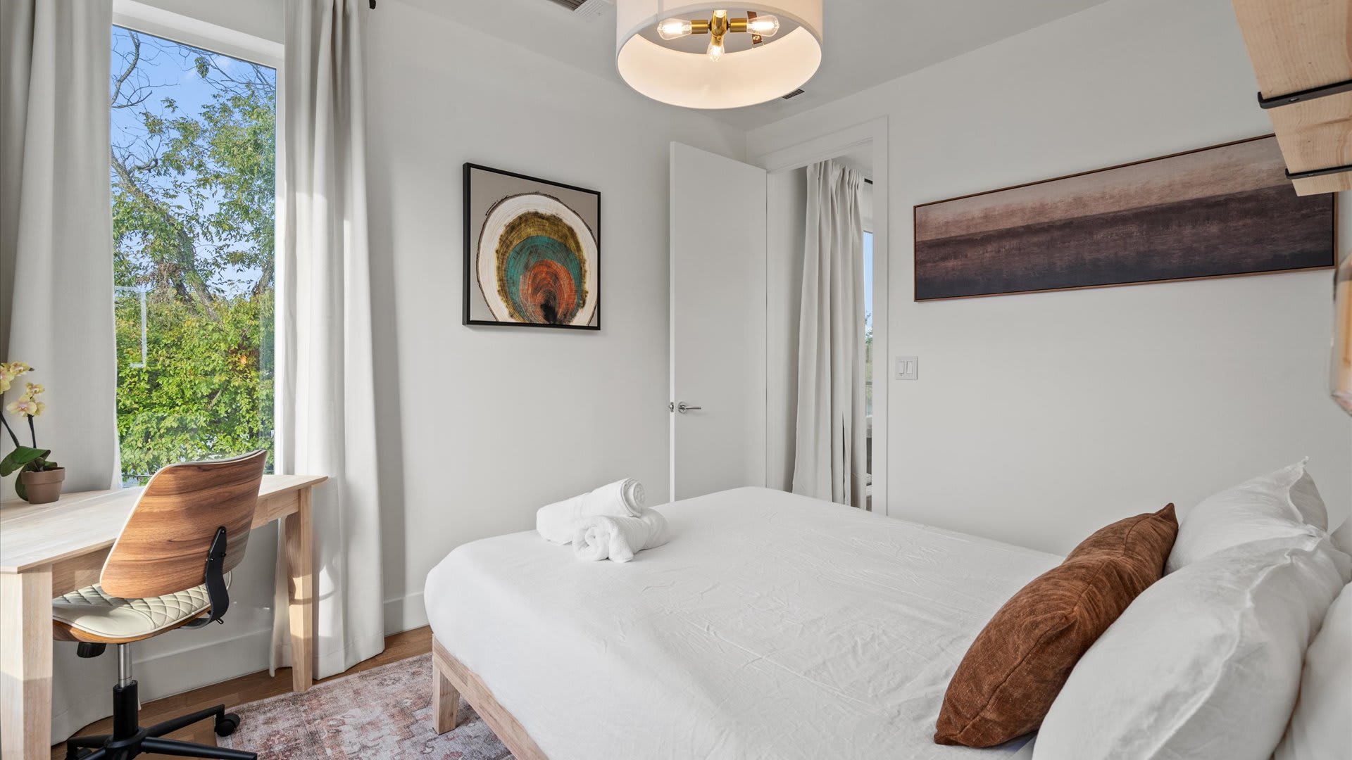 Bright bedroom with a single bed, white linens, a wooden desk with an office chair, and a large window offering a view of lush greenery. Artwork with concentric circles hangs above the bed.