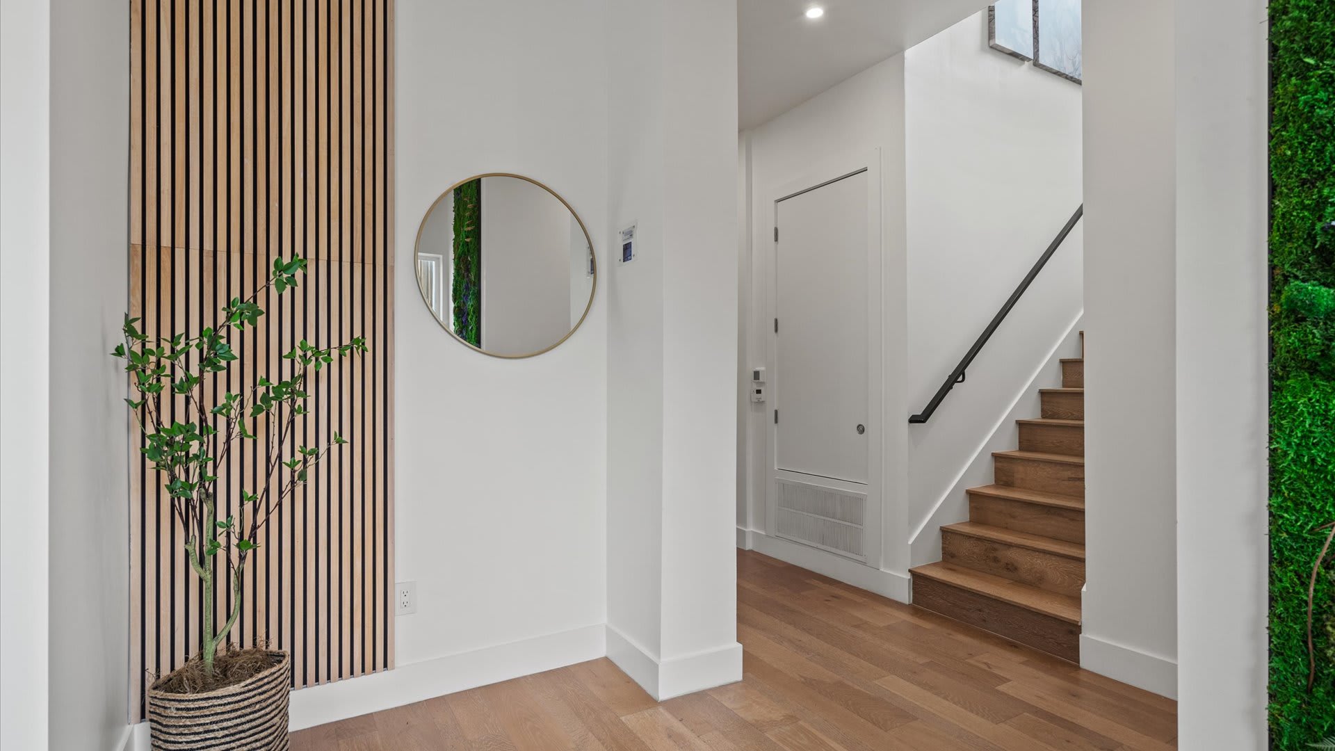 Bright and inviting foyer with light wood flooring, a striking vertical slatted wood accent wall, and a round gold-framed mirror. A staircase with a dark handrail leads to the upper level, complemented by a tall potted plant.
