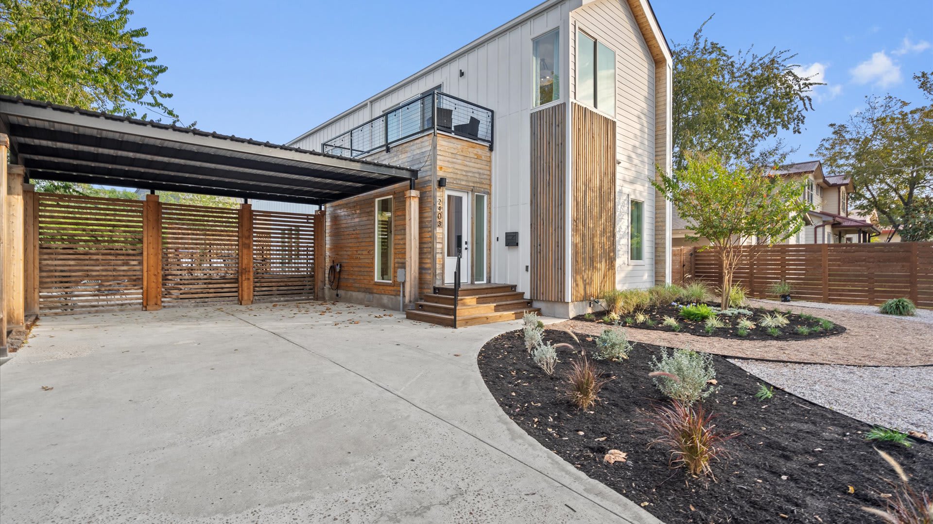 A wider view of the contemporary white and wood-sided house, showcasing a covered carport, a curved concrete driveway, and a meticulously landscaped front yard with gravel and dark mulch.