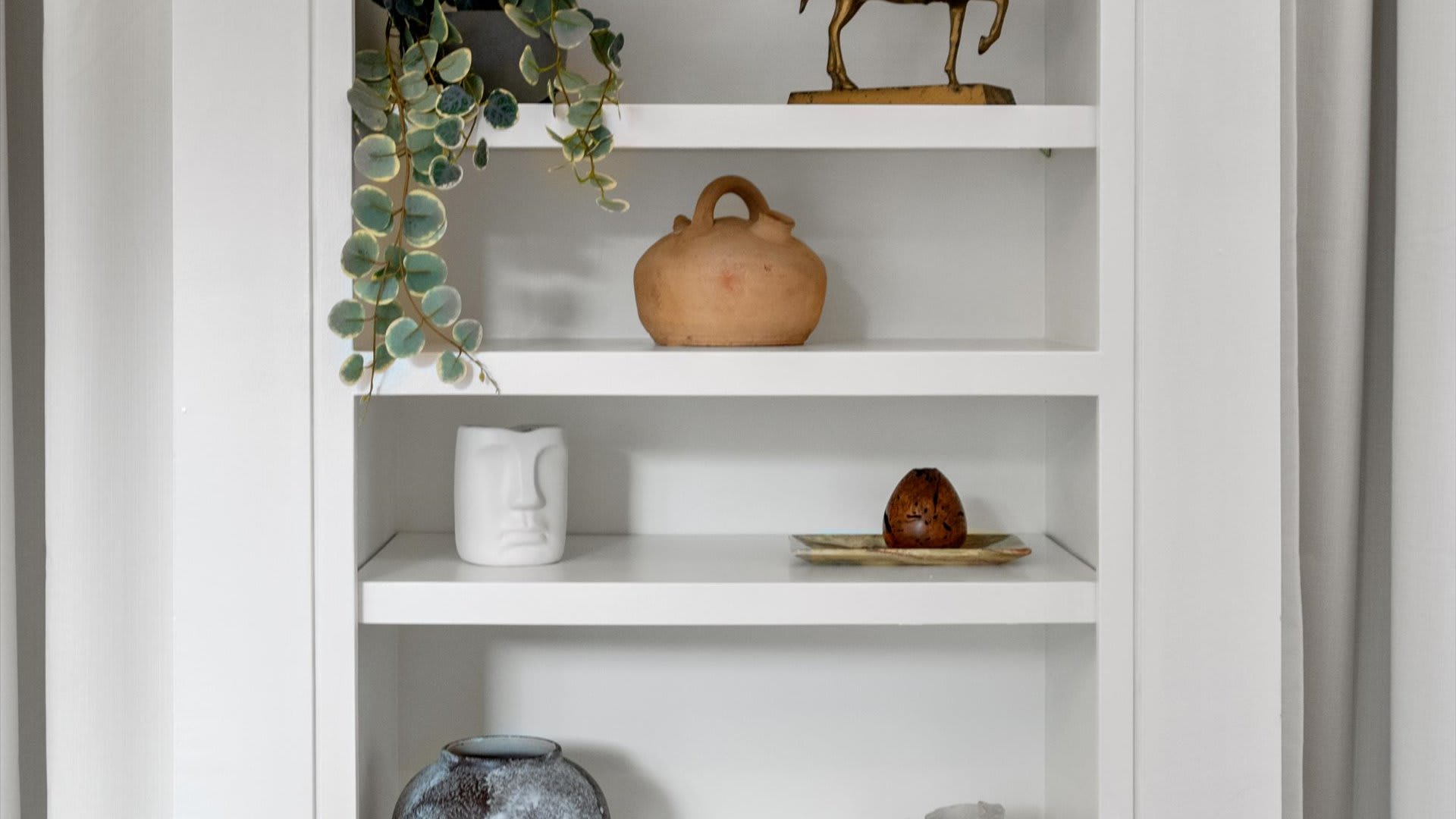 Close-up view of white built-in shelves, neatly displaying a variety of decorative items including a trailing potted plant, a rustic clay jug, a modern ceramic face vase, and other unique ornaments.