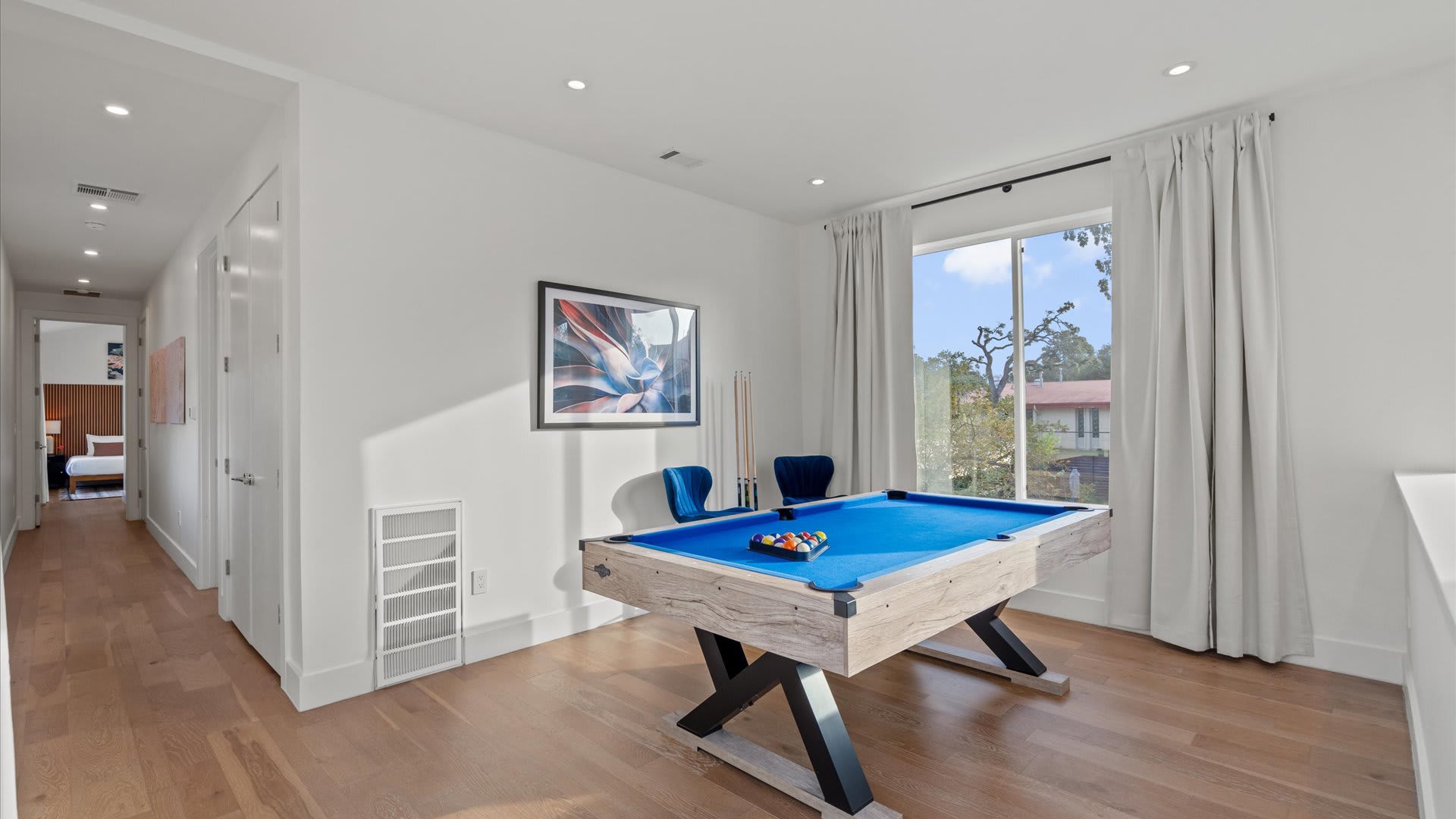Bright game room featuring a blue-felt pool table with a light wood and black base, accompanied by two blue velvet chairs. A large window with white curtains offers natural light, and a framed abstract artwork adorns the wall.