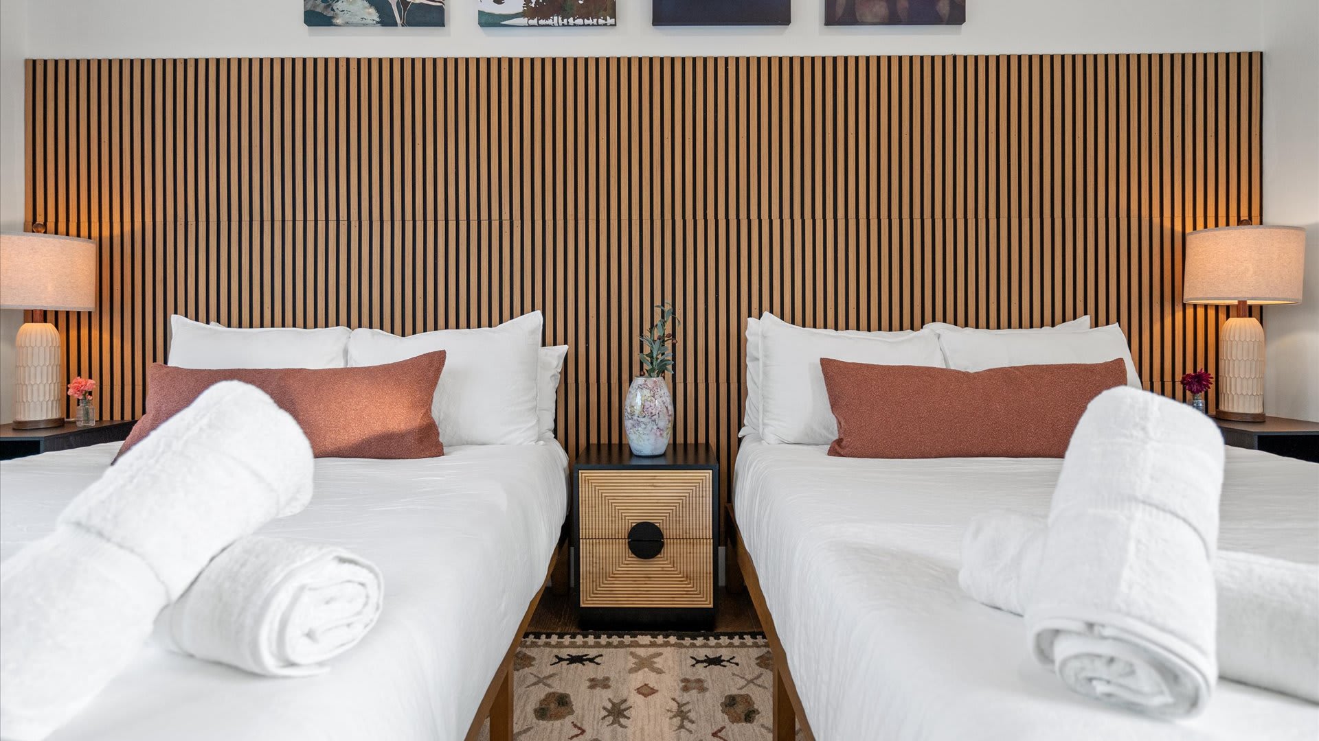 Close-up of the two beds in bedroom-3, showing white linens, rust-colored pillows, and a central wooden nightstand with a vase. The slatted wooden headboard spans the wall behind the beds.