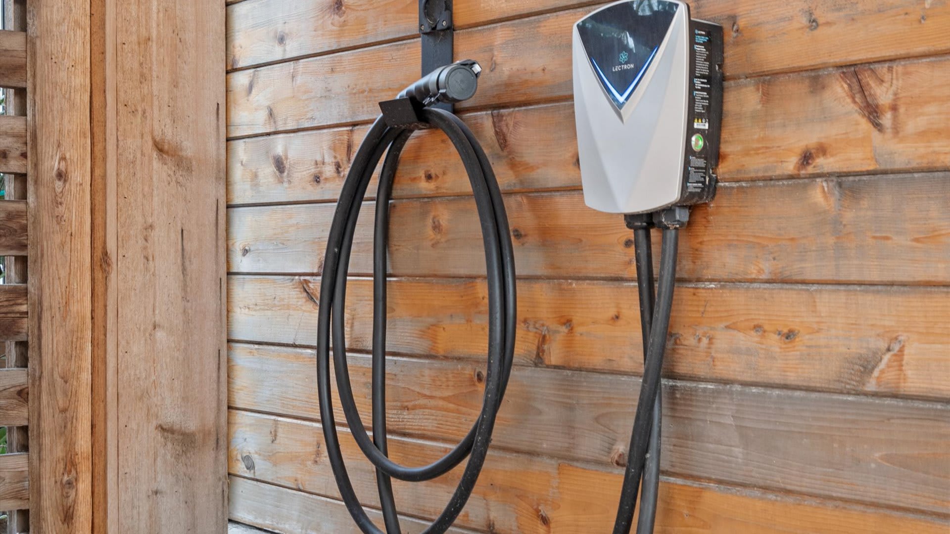 A white and gray electric vehicle charging station mounted on a rustic wooden plank wall, with its coiled black cable neatly hung beside it.