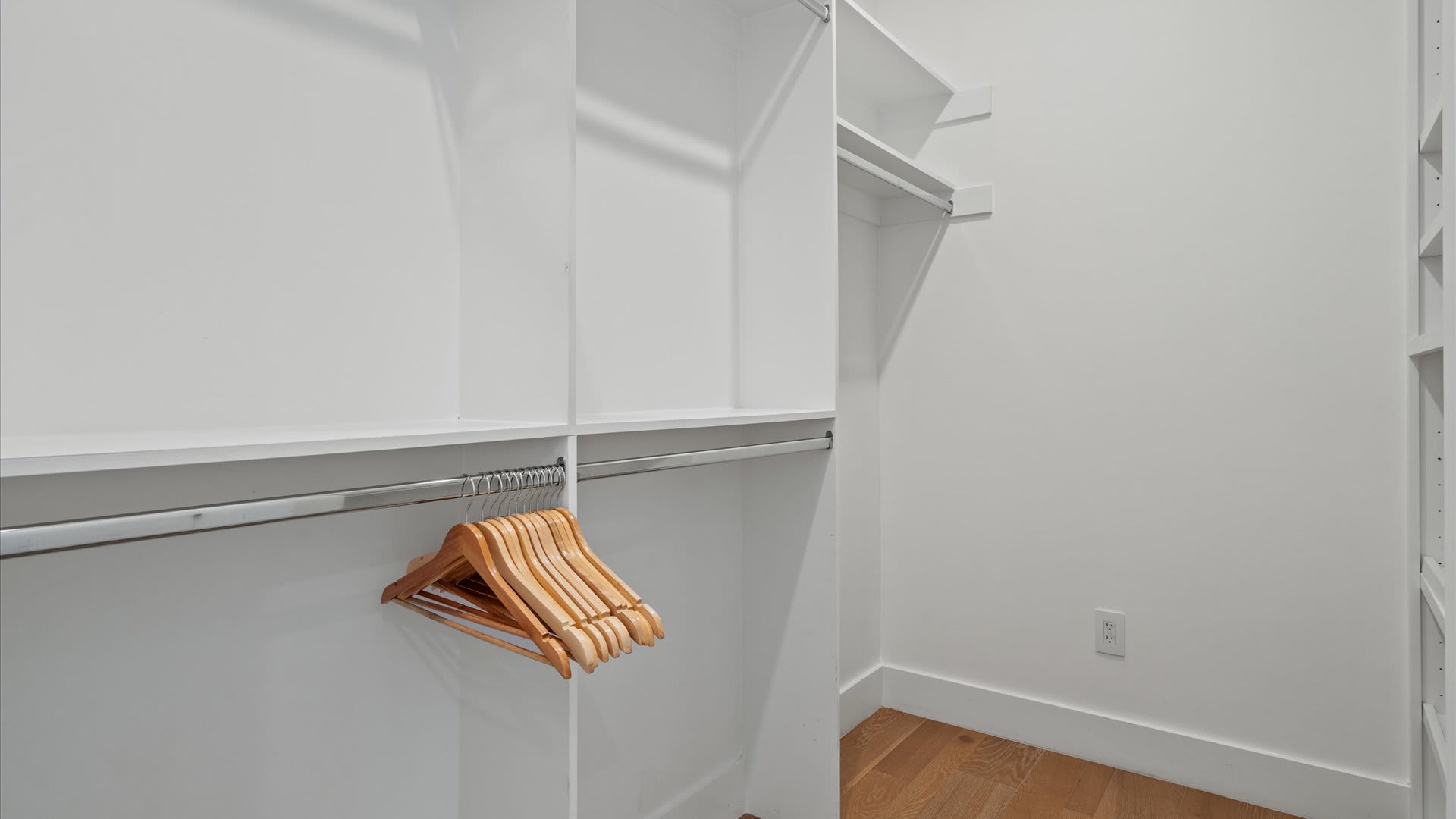 A clean, empty walk-in closet with white shelving units, a metal hanging rod, and a stack of wooden hangers, set against light hardwood flooring.