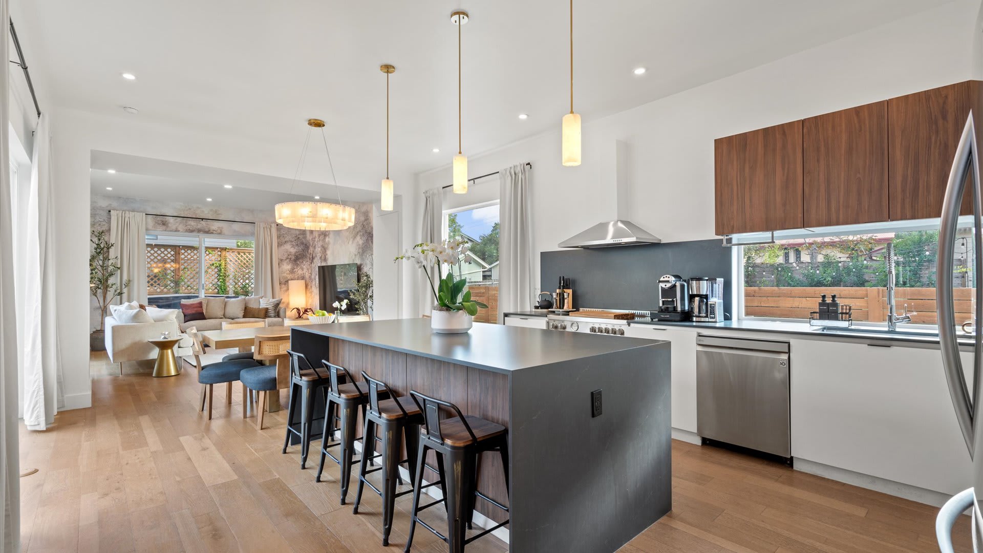 Modern kitchen featuring a large dark island with three industrial-style bar stools, stainless steel appliances, and warm wood cabinetry. Pendant lights hang above the island, and the open space flows into the dining and living areas.