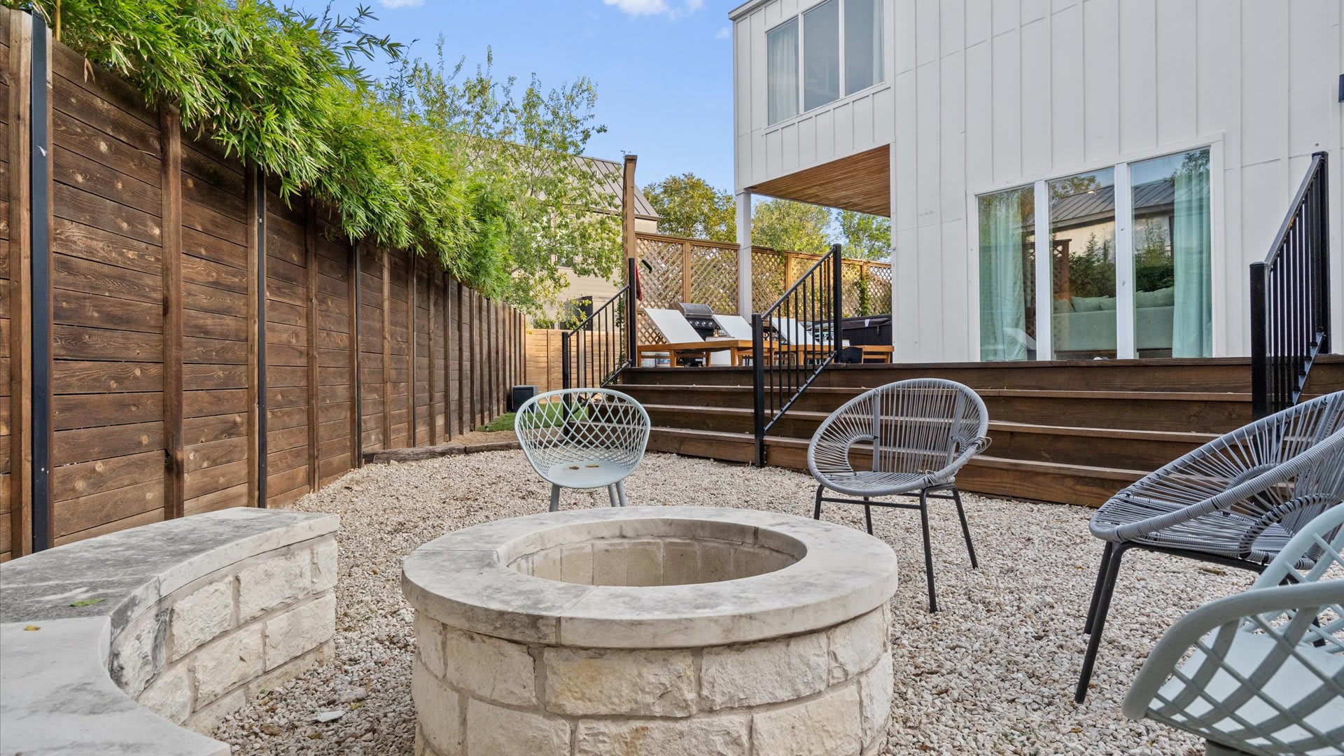 A close-up of the stone fire pit in the gravel backyard, featuring a circular stone ring and a curved stone bench, complemented by modern wire-frame chairs. A tall wooden fence borders the property, ensuring privacy.