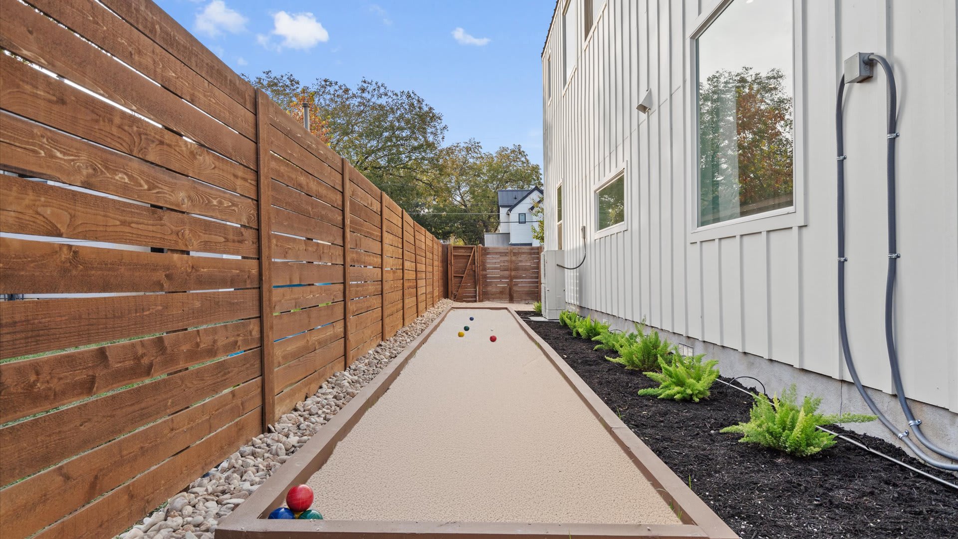 A narrow side yard features a long, sandy bocce ball court bordered by dark mulch and small green plants. A tall wooden fence runs along one side, and the white exterior of the house is visible on the other.