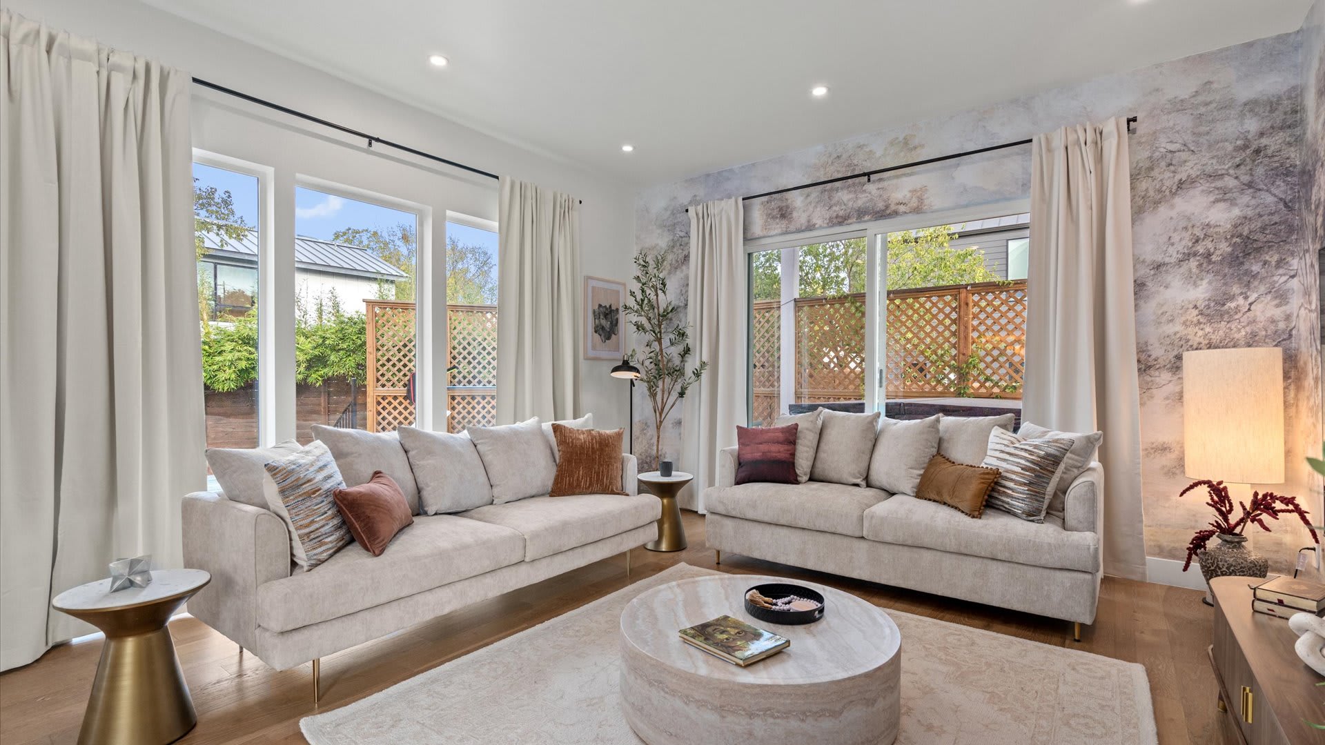 Modern living room with two light-colored sofas adorned with throw pillows, centered around a round marble coffee table. Large windows with white drapes provide ample light, and an accent wall features a subtle, artistic mural wallpaper.