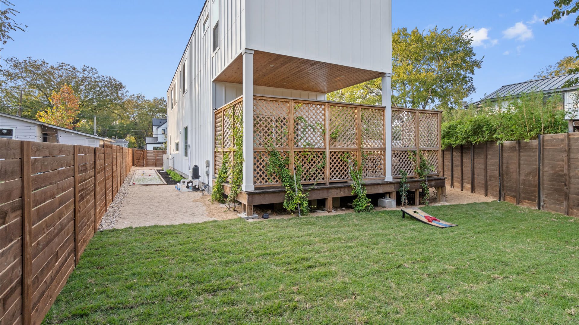 Spacious backyard featuring a well-maintained grassy lawn, a sand bocce ball court, and a raised wooden deck with lattice privacy screens. A cornhole game board rests on the grass, and a tall white house exterior is visible.