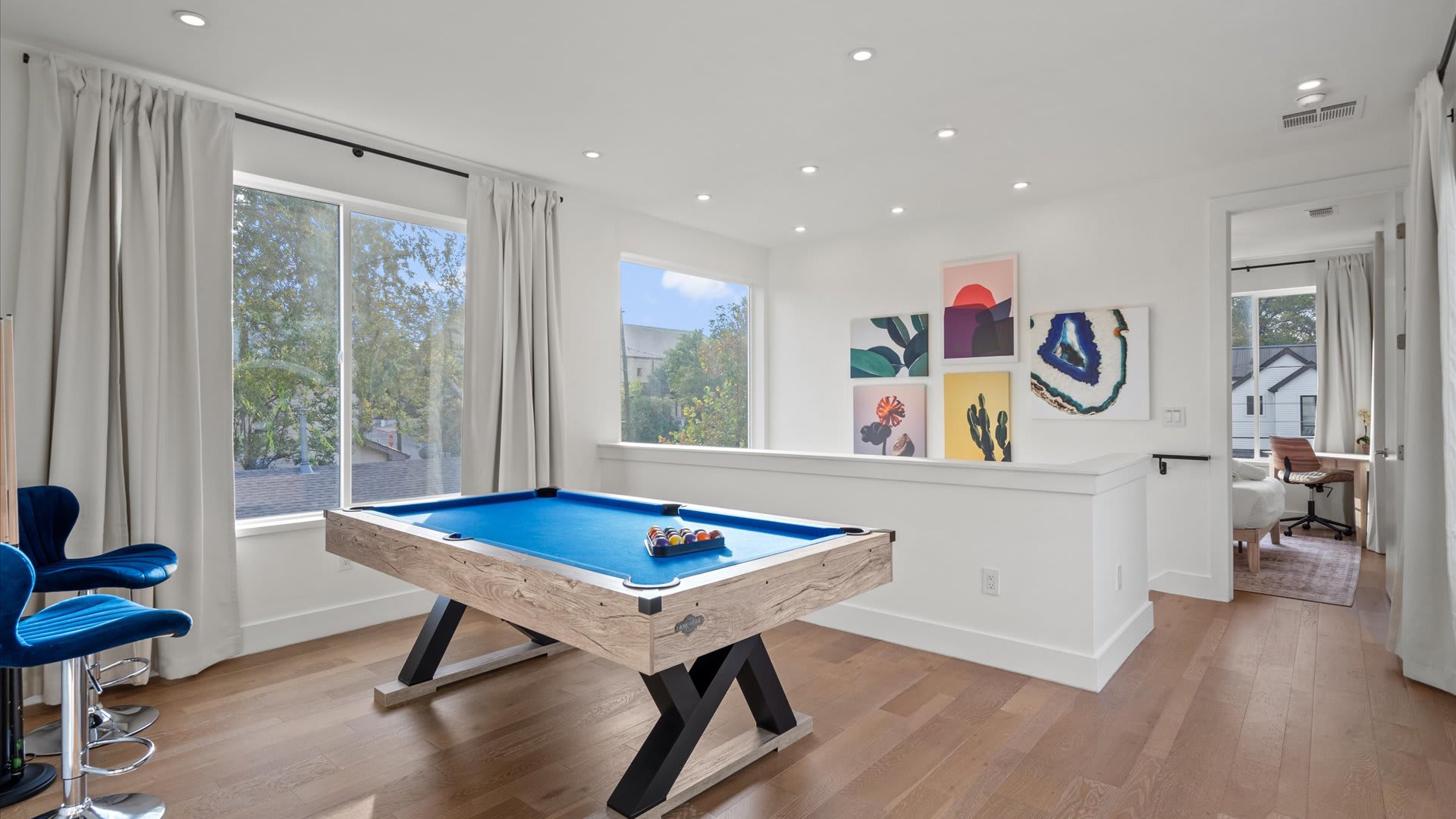 A bright game room on an upper floor features a modern wooden pool table with a vibrant blue felt surface, surrounded by large windows with white curtains. Colorful abstract art adorns the white walls, and a small office nook is visible in the background.