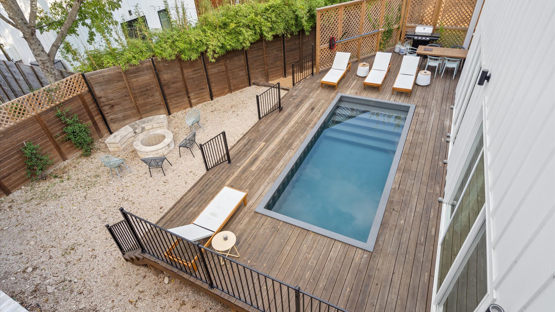 An aerial view of the backyard reveals a rectangular plunge pool set into a wooden deck, surrounded by lounge chairs. Beyond the deck, a gravel yard features a stone fire pit with seating, all enclosed by a tall wooden fence.