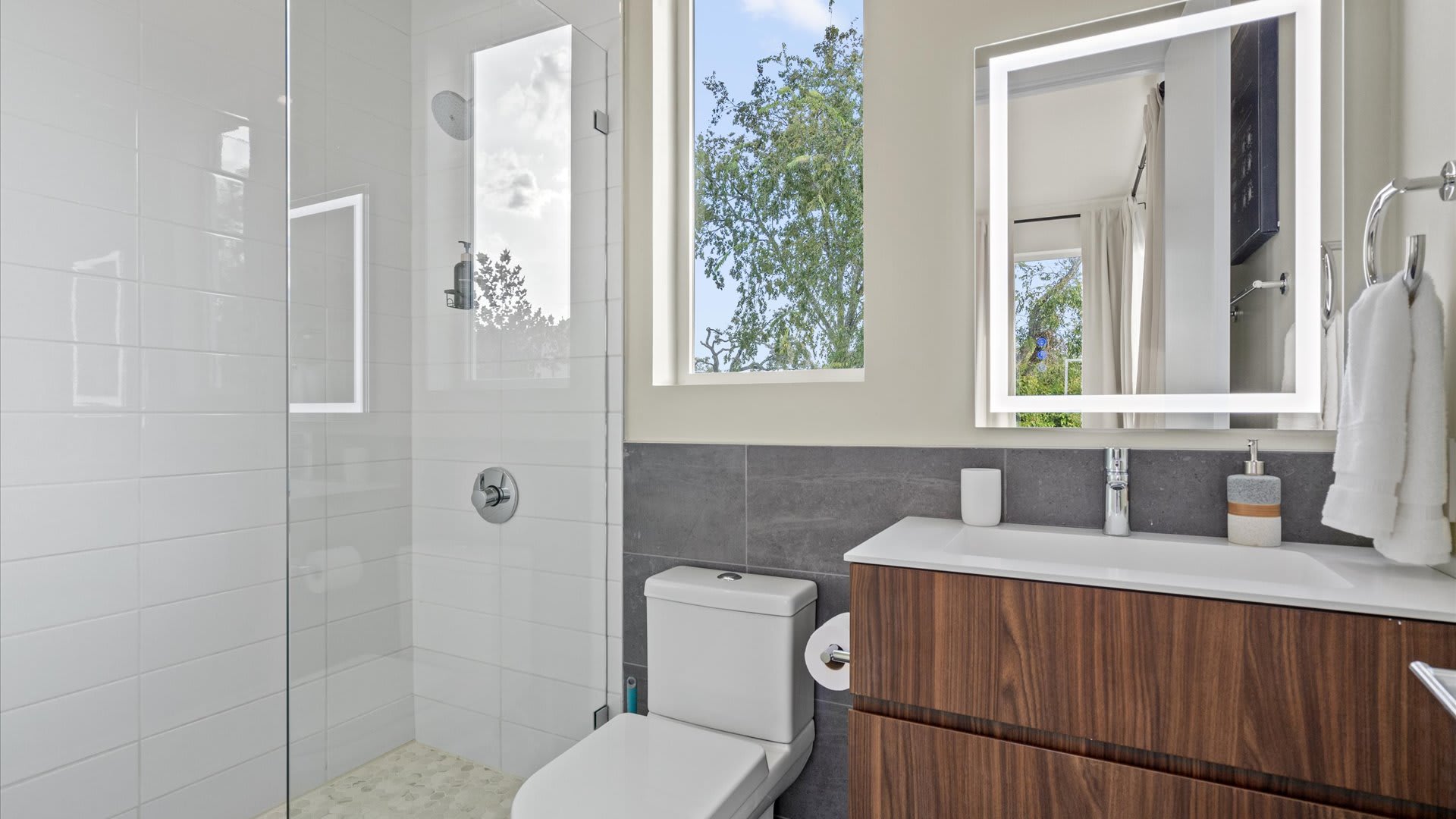 Modern bathroom featuring a glass-enclosed shower with white subway tiles, a toilet, and a dark wood vanity with a white sink and an illuminated mirror. A window shows outdoor greenery.
