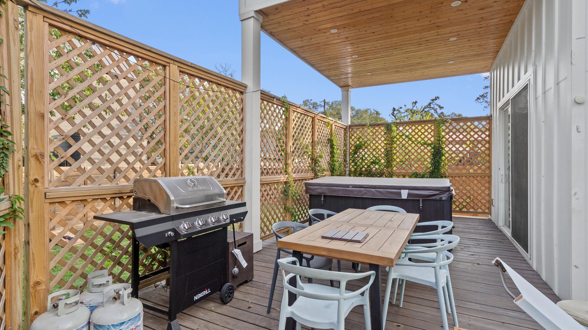 A covered outdoor patio features a wooden dining table with light blue chairs, a stainless steel BBQ grill, and a dark hot tub. Wooden lattice screens provide privacy and a cozy atmosphere on the wooden deck.