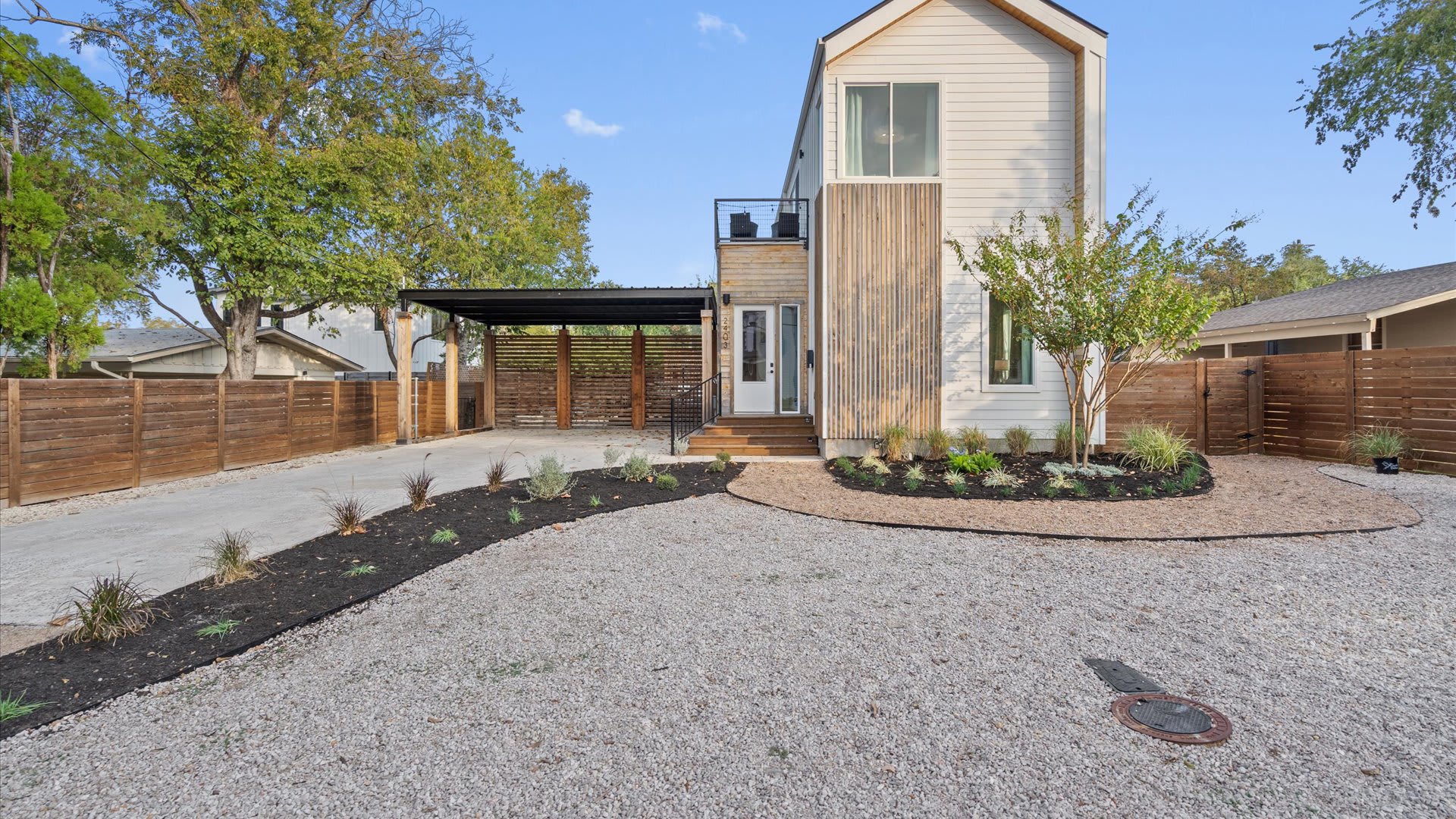 The modern white and wood-paneled exterior of the house, featuring a covered carport, a concrete driveway, and a landscaped front yard with gravel and low-maintenance plants.