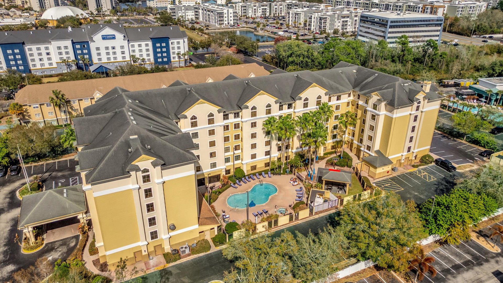 Expansive aerial view of a large hotel complex with a central courtyard featuring a swimming pool and hot tub, surrounded by palm trees and lounge chairs. Adjacent buildings and parking areas are also visible.
