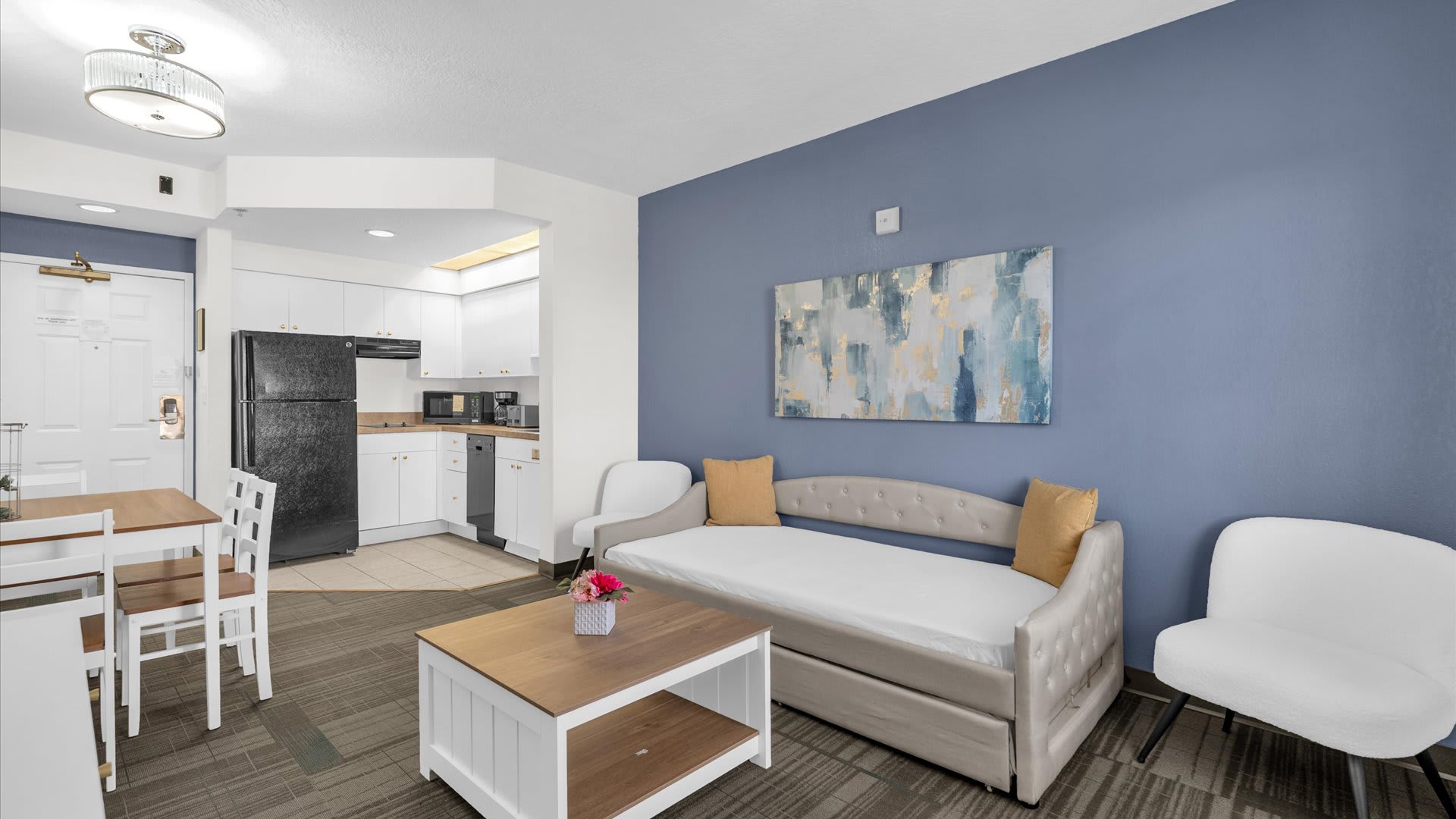 Open-concept living space with a blue accent wall, a light gray daybed, and a white and wood coffee table, leading into a compact kitchen with white cabinetry and black appliances.