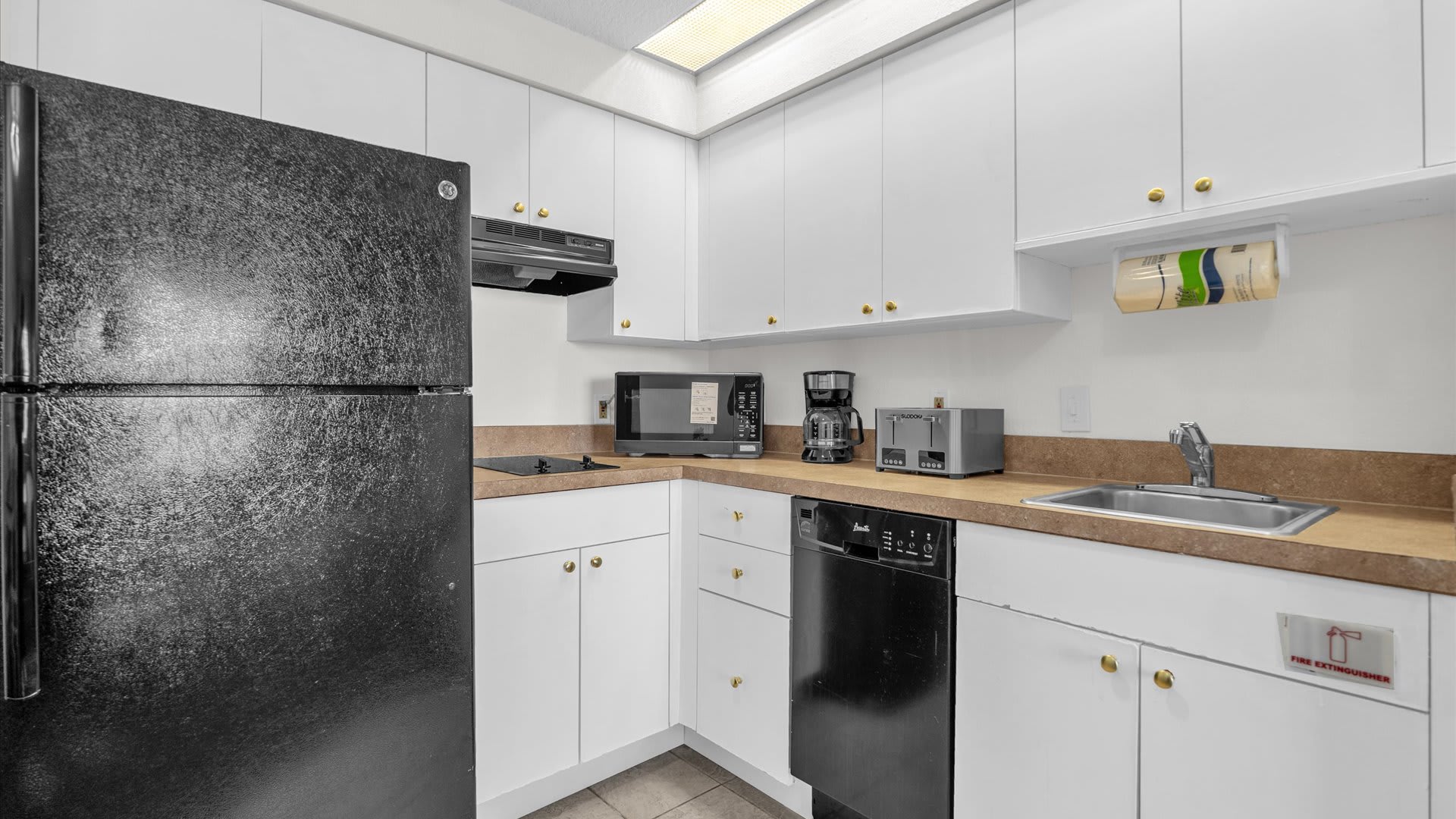 Well-equipped kitchen counter with a black dishwasher, stainless steel sink, toaster, microwave, and coffee maker, set against white cabinets and a light brown countertop.