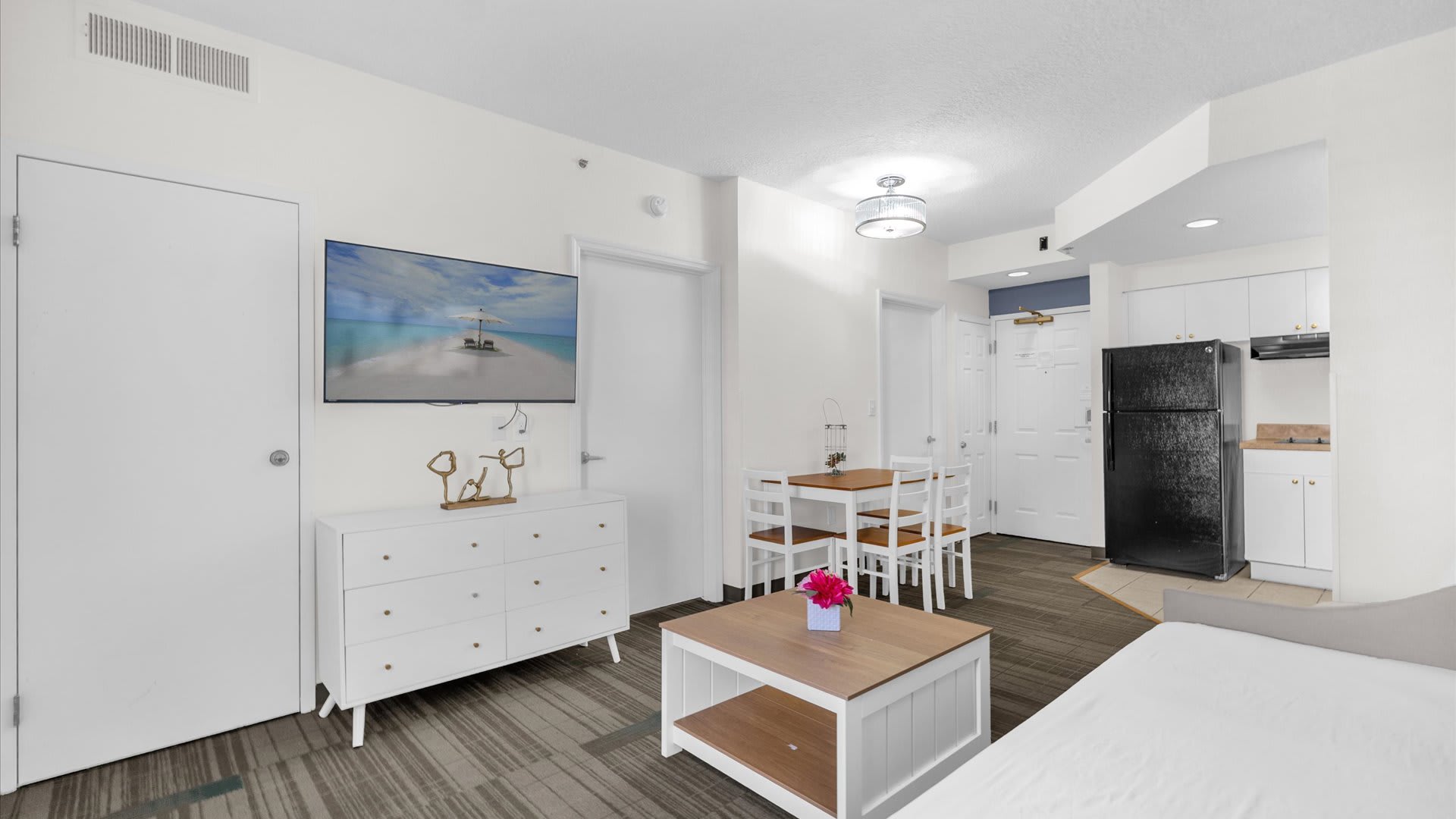 Modern open-plan living and dining area with a wall-mounted TV above a white dresser, a wooden dining table with white chairs, and a coffee table, leading to a daybed in the background.