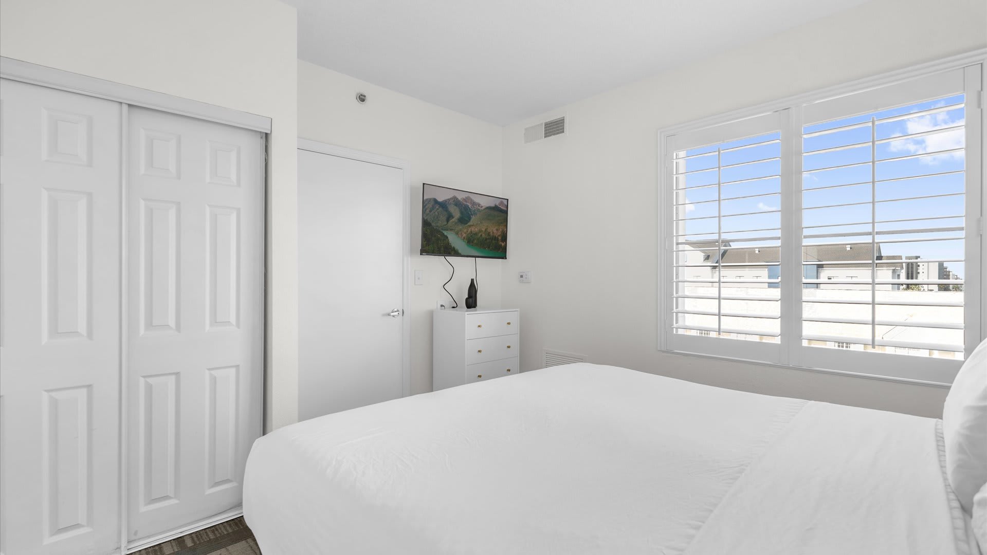 Bright bedroom featuring a white bed with crisp linens, a large white closet with bi-fold doors, a wall-mounted TV, and a window with white plantation shutters.