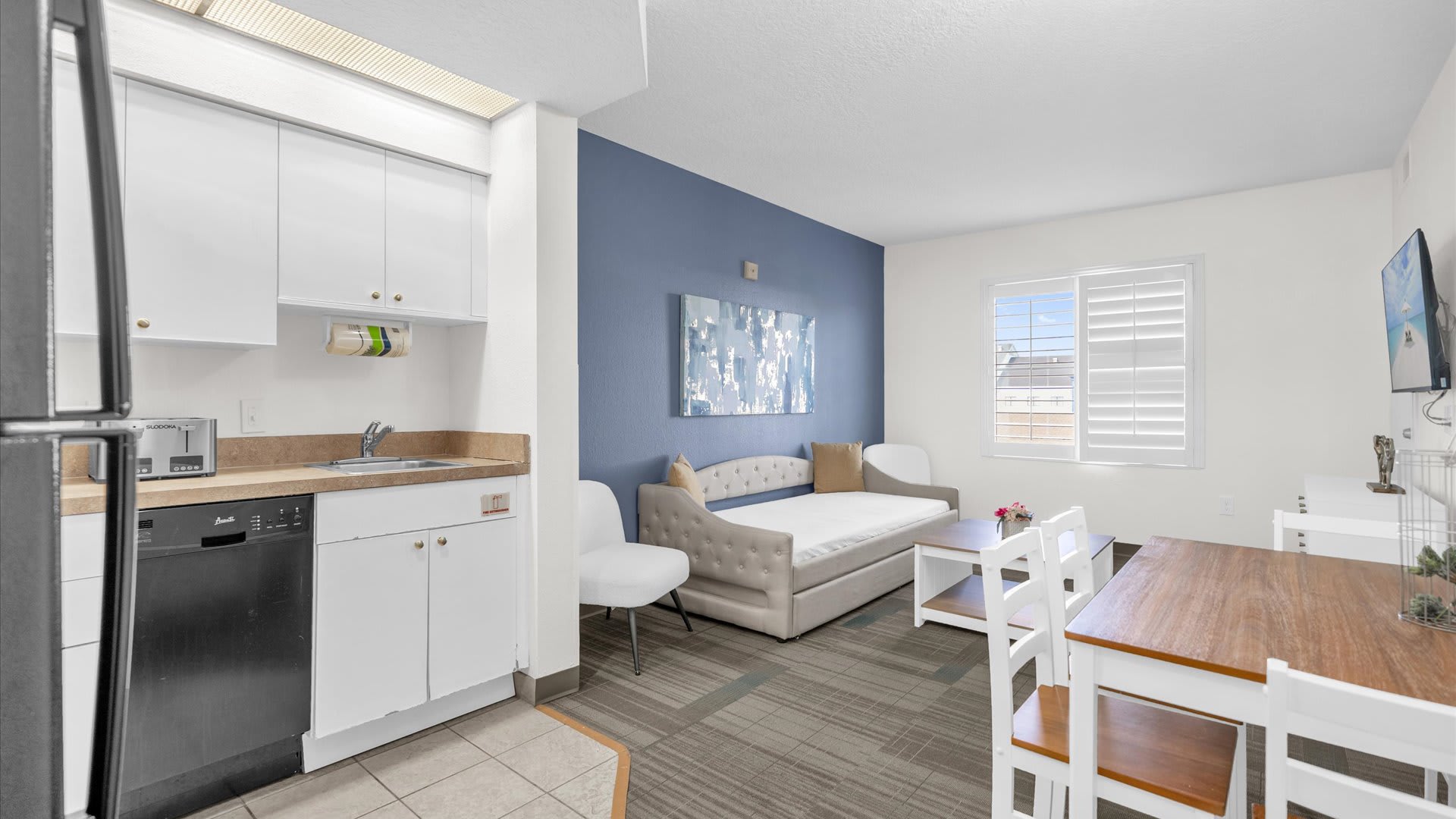 Bright living area with a blue accent wall and a daybed, adjacent to a compact kitchen featuring white cabinets, a black dishwasher, and a stainless steel sink, all under a fluorescent light fixture.