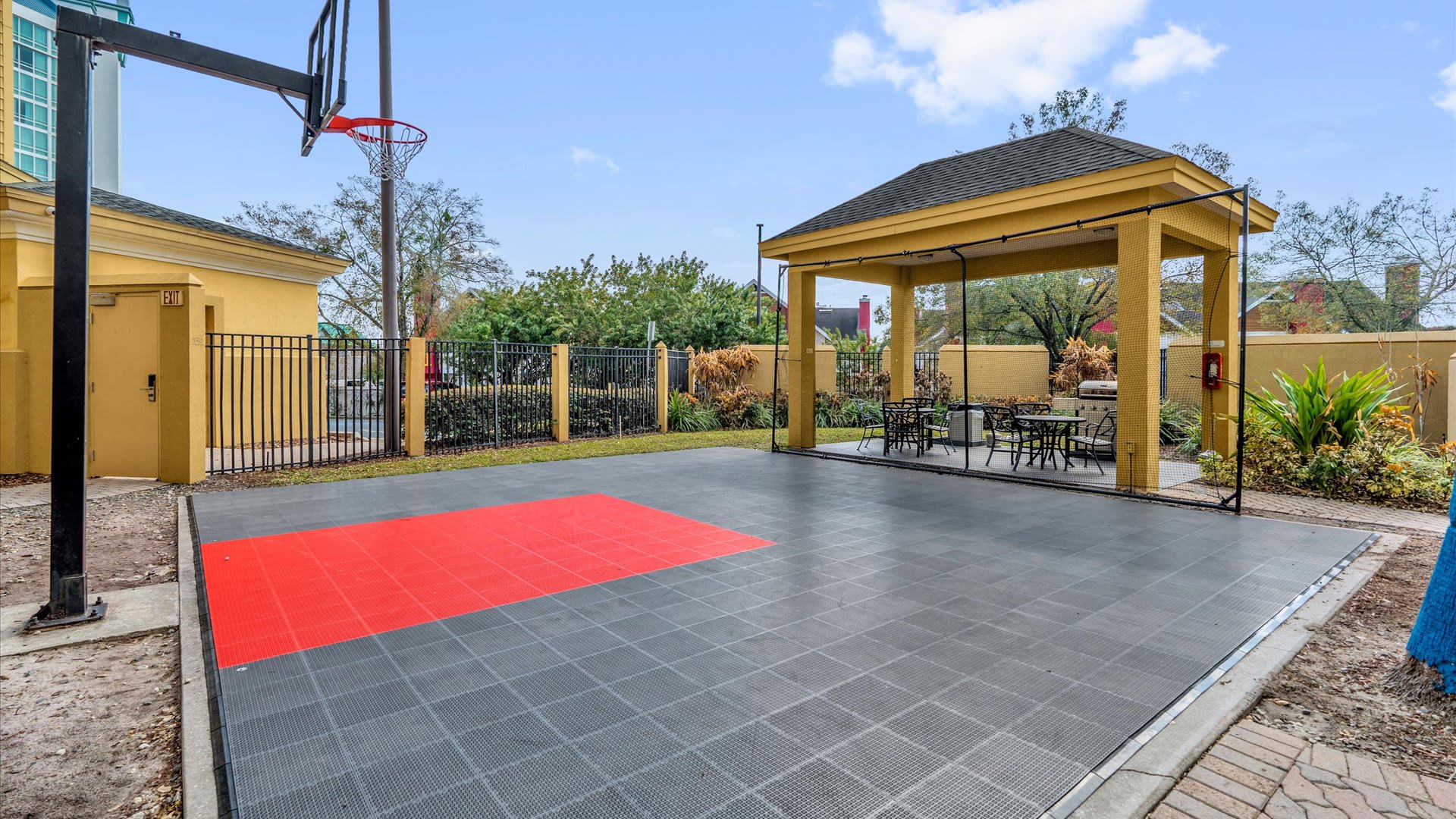 Outdoor recreational area featuring a basketball court with a red and gray tiled surface, a basketball hoop, and a yellow covered gazebo with outdoor seating for relaxation.