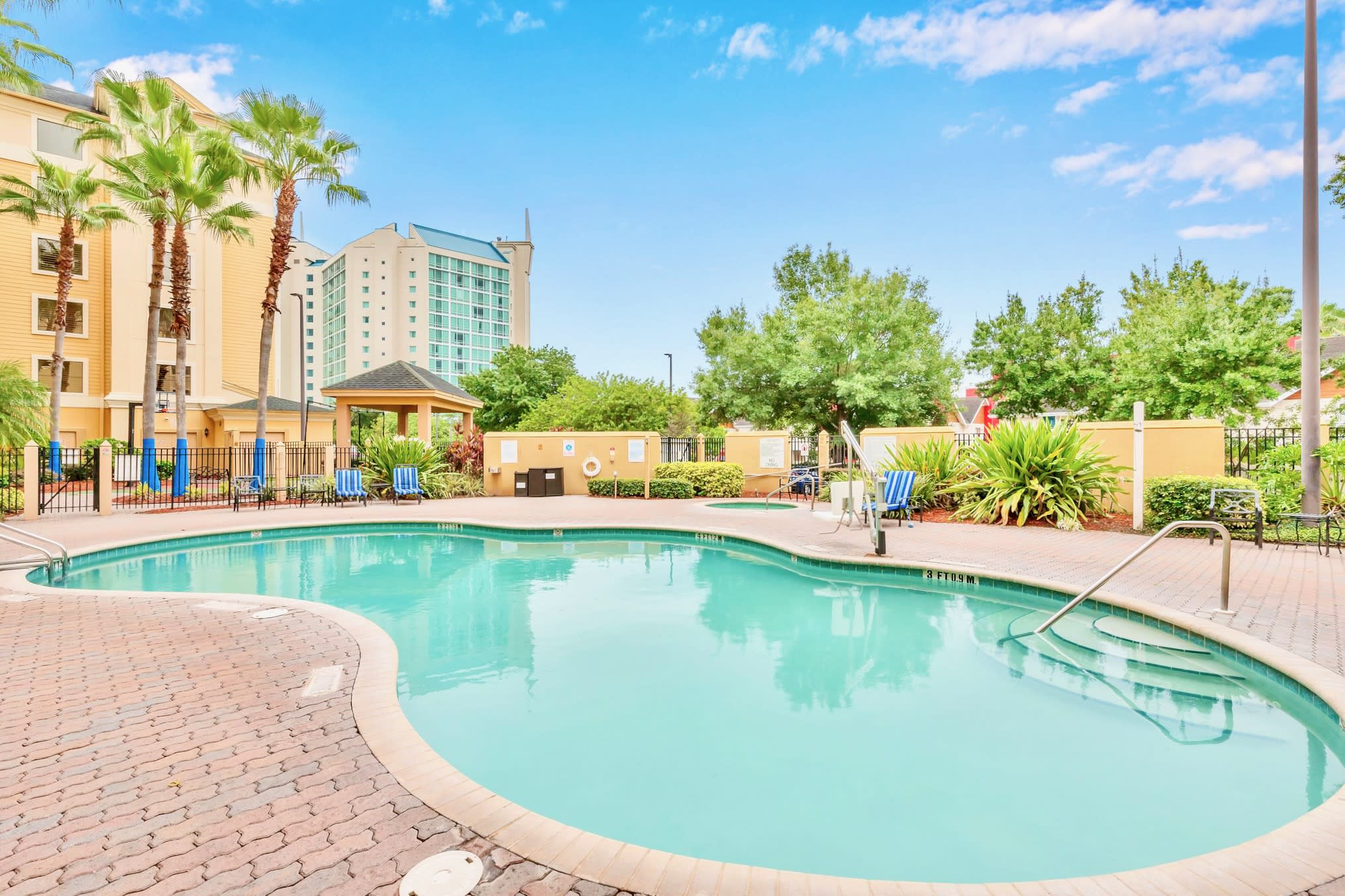 A large, irregularly shaped swimming pool with light blue water is surrounded by a brick paver deck, with palm trees and a multi-story building in the background under a bright blue sky.