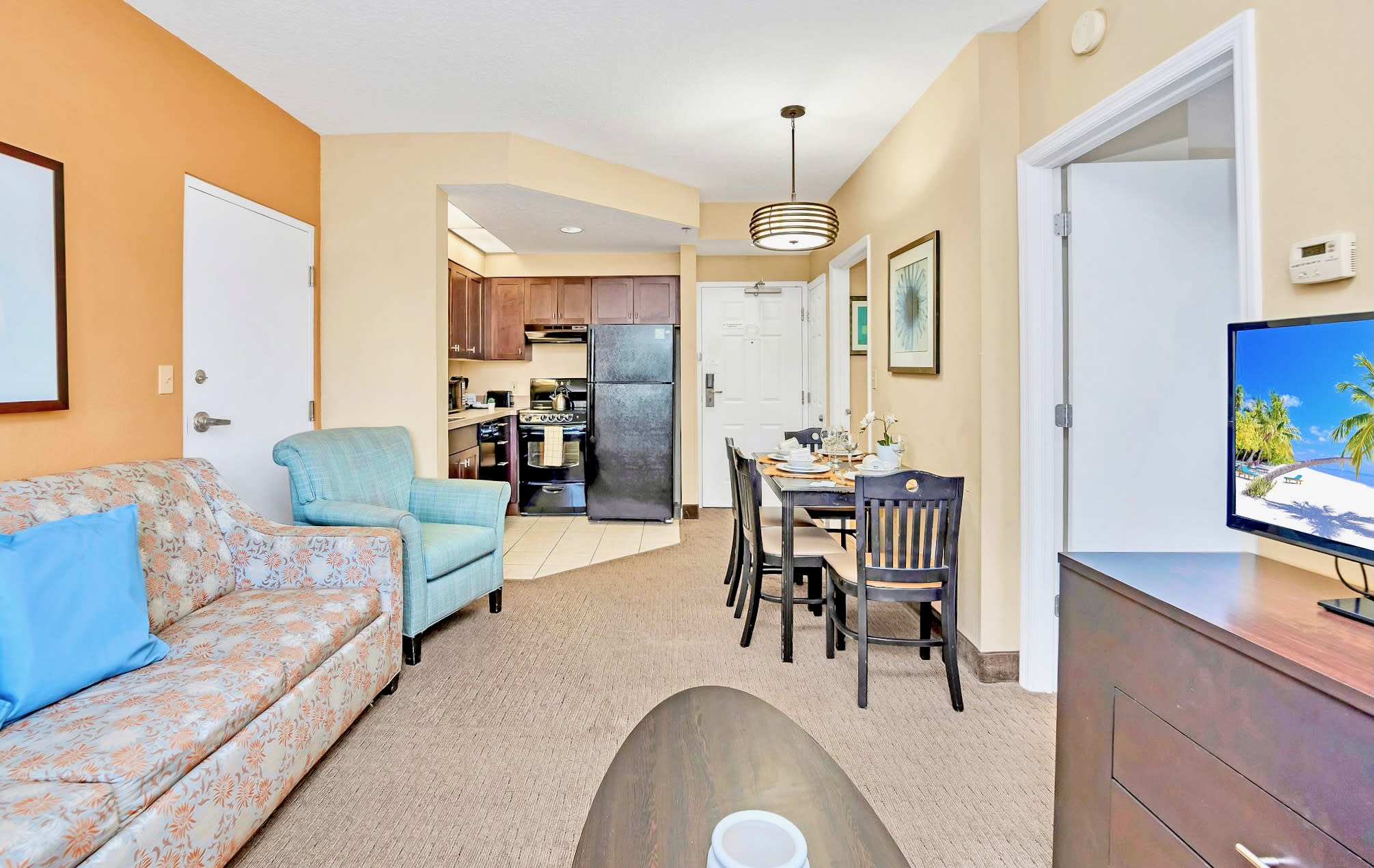 A wider view of the open-concept living and dining area, showing the dark wood dining table, floral sofa, and light blue armchair, with a partial view of the adjacent kitchen and a TV on a dark dresser.