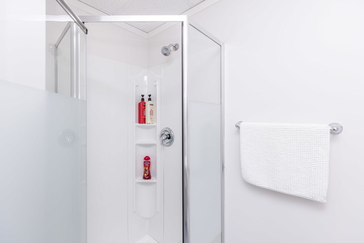 A bright, white shower enclosure with clear glass panels and a frosted glass wall, featuring a built-in corner shelf holding shampoo and conditioner bottles. A white textured towel hangs on a chrome bar outside the shower.