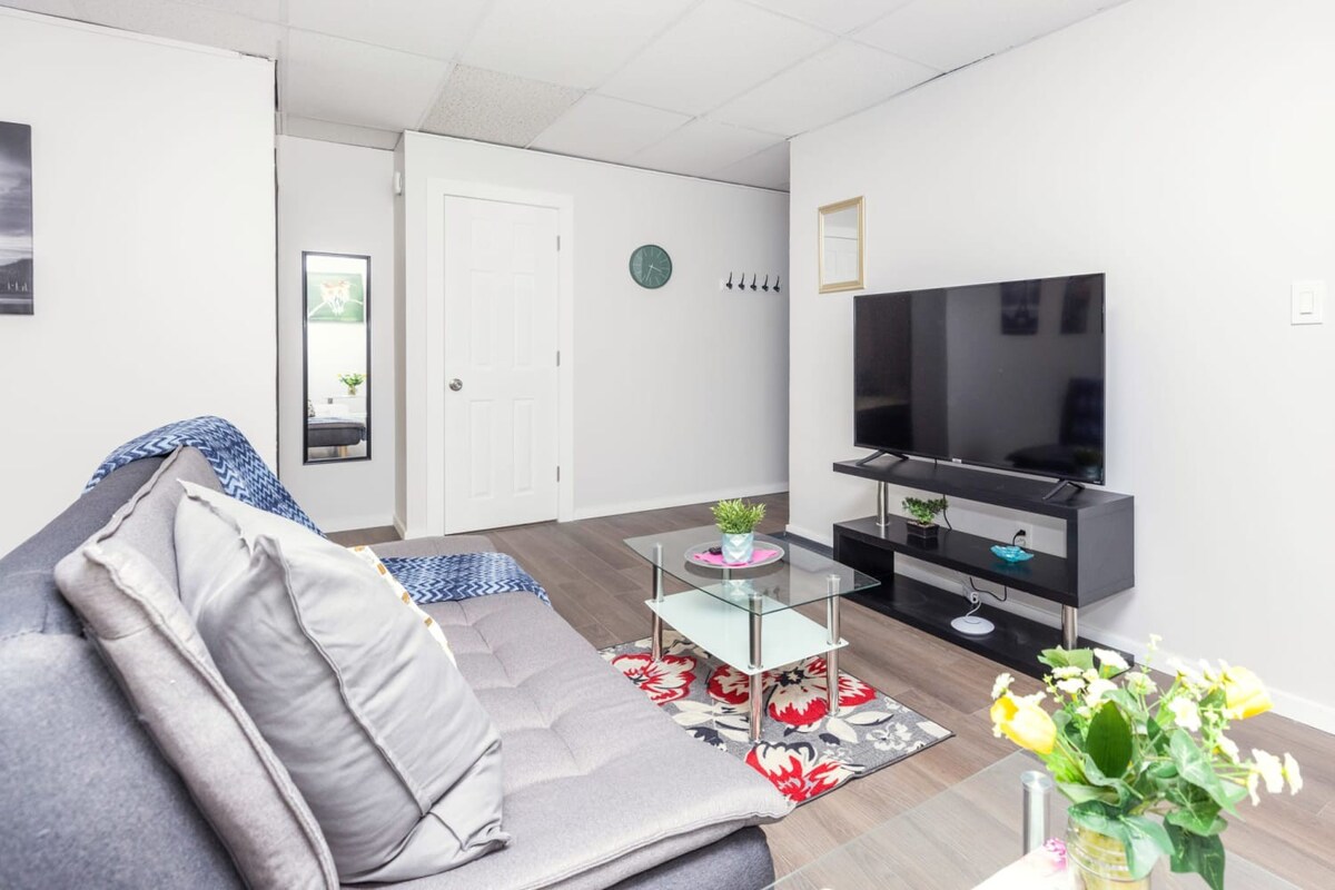 View of the living room from the sofa, showcasing a wall-mounted flat-screen TV on a black stand, a glass coffee table with a small plant, and a white door leading to another room.
