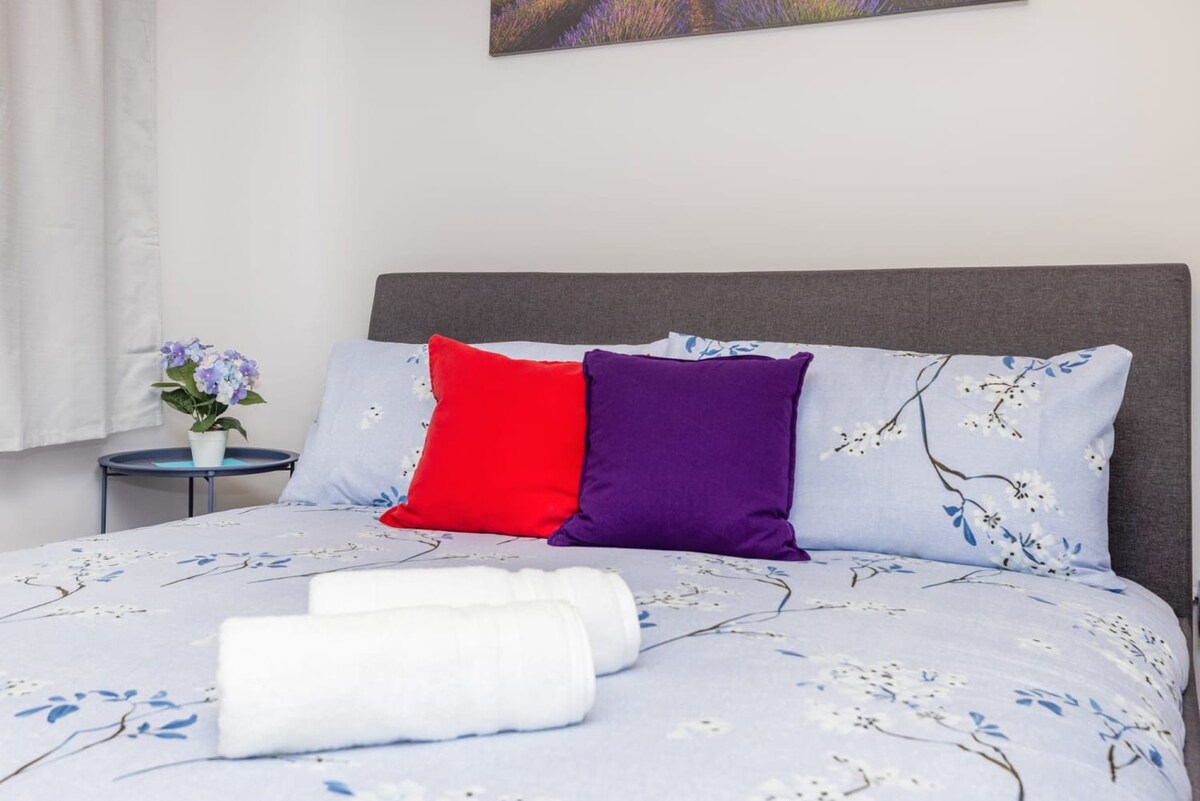 Close-up of the gray upholstered bed with a light blue floral duvet, neatly rolled white towels, and vibrant red and purple accent pillows, next to a small blue bedside table with a plant.