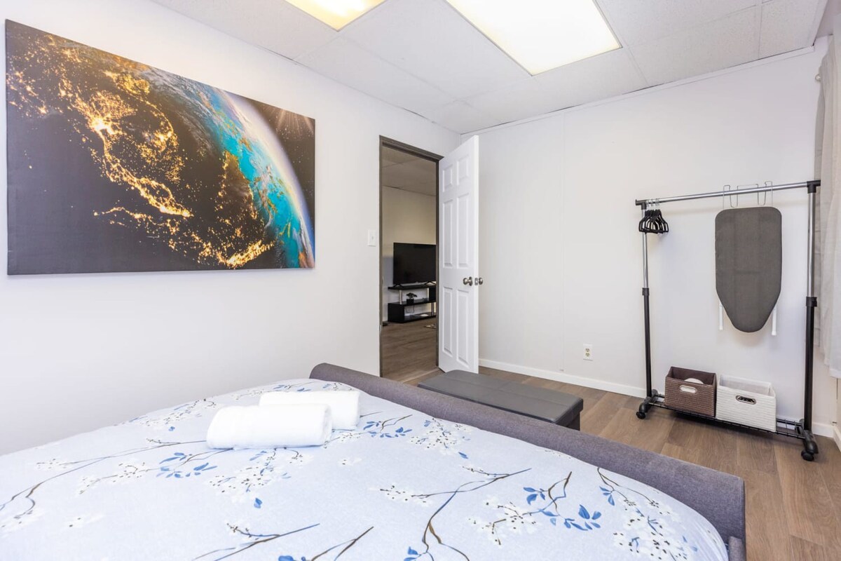 A modern bedroom with light gray walls, a bed featuring a light blue floral duvet, and a large artwork depicting Earth from space. A black clothing rack with an ironing board stands in the corner, and a doorway leads to another room with a TV.