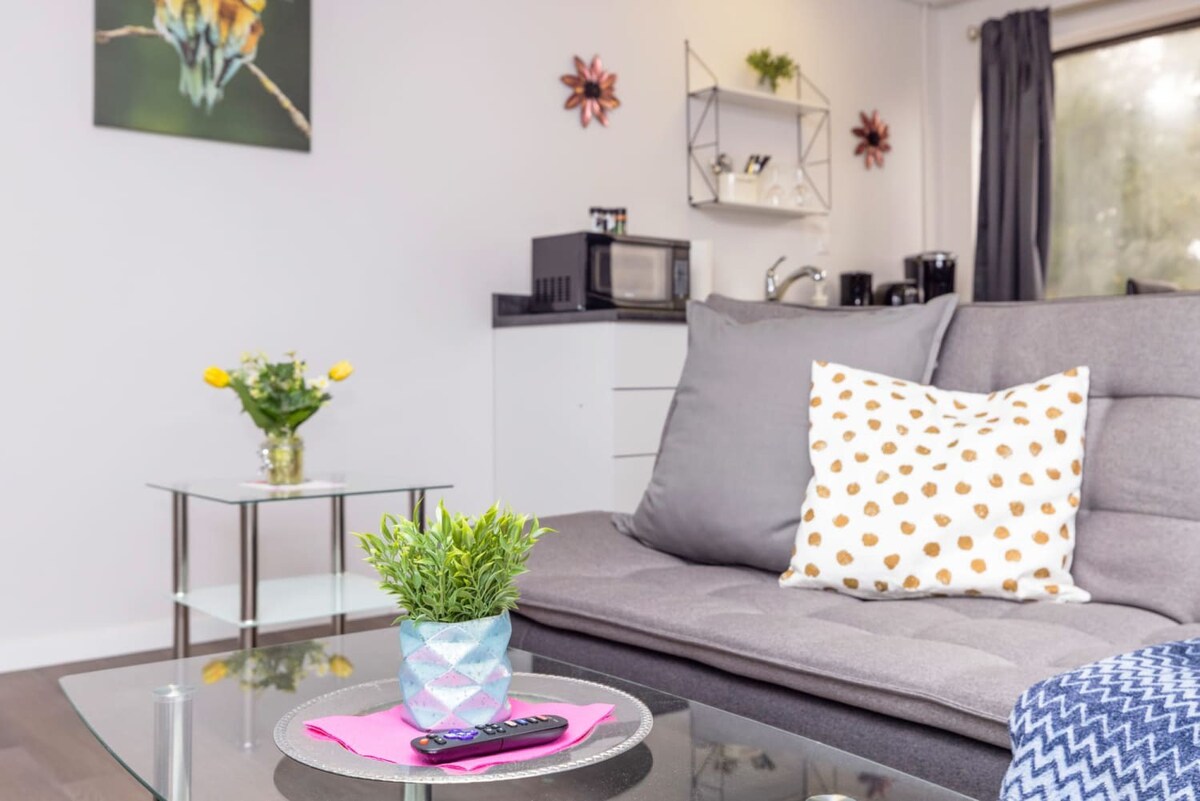 Close-up of the gray futon sofa adorned with decorative pillows and a blue throw blanket, a glass coffee table holding a small potted plant and remote, and a side table with flowers.