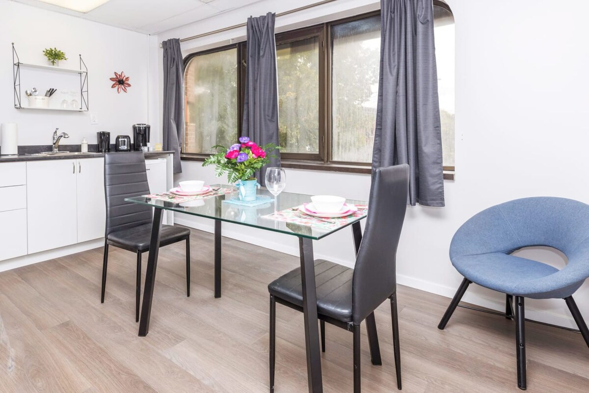 Modern dining area with a glass-top table and two black chairs, positioned next to a compact kitchen counter and a blue accent chair, with large windows providing natural light.