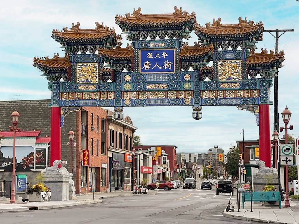 A vibrant street scene featuring a large, ornate traditional Chinese gate with intricate blue, gold, and red details, marking the entrance to Chinatown. The street below is lined with buildings and cars under a clear blue sky.