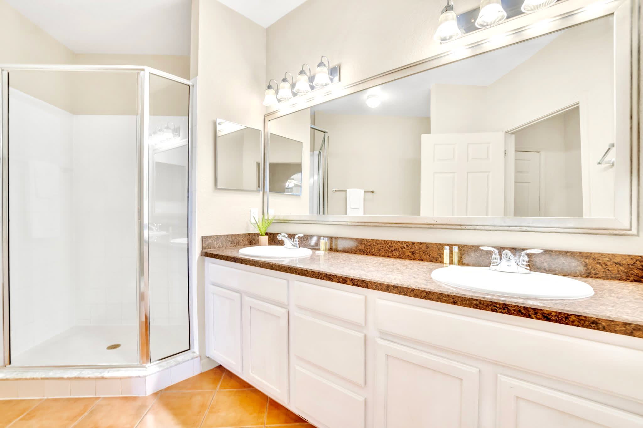 Bathroom vanity with double sinks, a large mirror, and white cabinets. A glass-enclosed shower is visible to the left, and the floor is tiled, providing a functional and bright space.