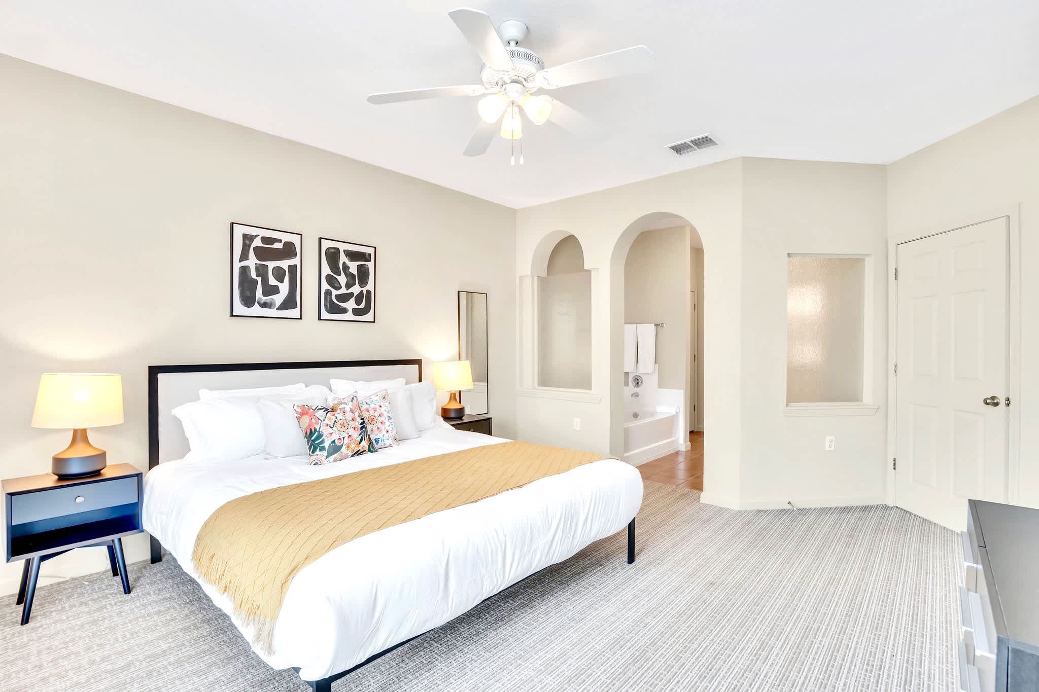 Spacious bedroom with a king-sized bed, crisp white bedding, and a mustard yellow throw. Two black nightstands with lamps flank the bed, and abstract art hangs above the headboard, all on a light carpeted floor.