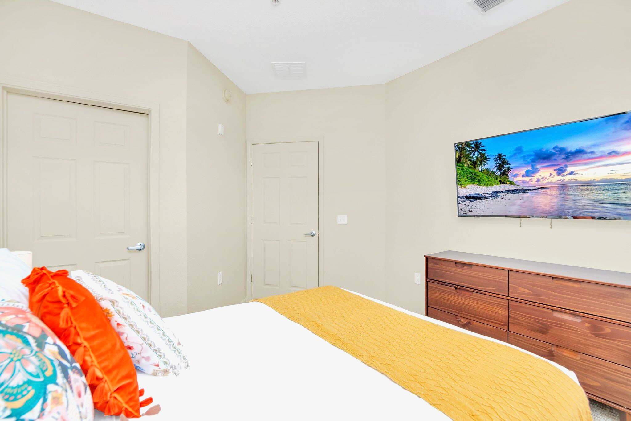 This bedroom view shows a wall-mounted TV above a dark wood dresser, with two light-colored doors visible in the background. The bed, partially visible, has white linens and a yellow throw.