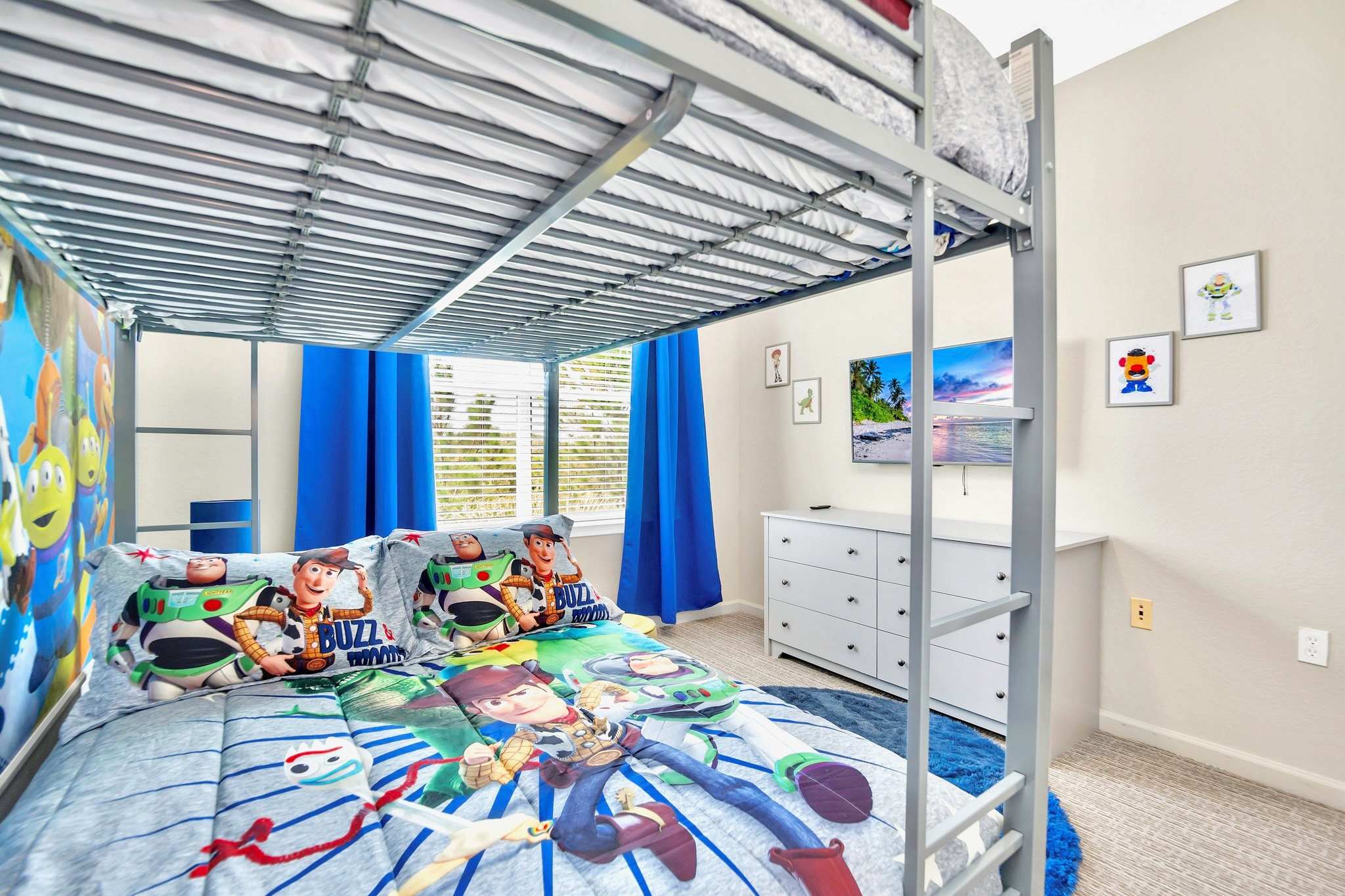 This view of the kids' bedroom shows the metal bunk bed with Toy Story bedding, a wall-mounted TV above a white dresser, and a window with blinds and blue curtains. Small framed artwork adorns the wall.