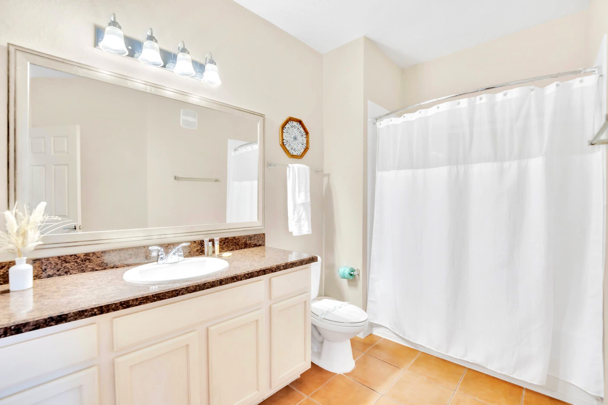 A bright bathroom features a single vanity with a granite countertop, a large mirror, and a shower/tub combo with a white curtain. The floor is tiled in a warm, earthy tone.