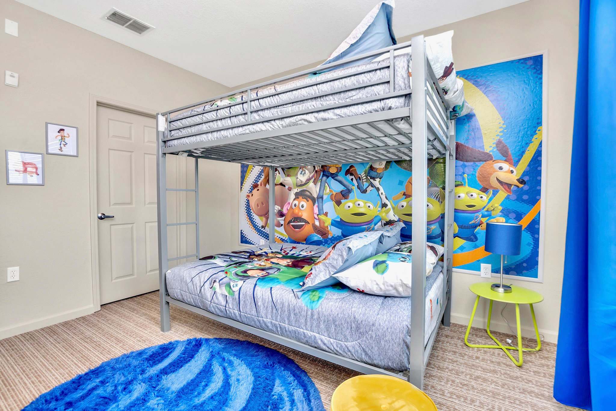 A vibrant kids' bedroom features a metal bunk bed with Toy Story themed bedding and a large wall mural. A small green side table with a blue lamp sits next to the bed, and a blue rug adds a pop of color to the carpeted floor.