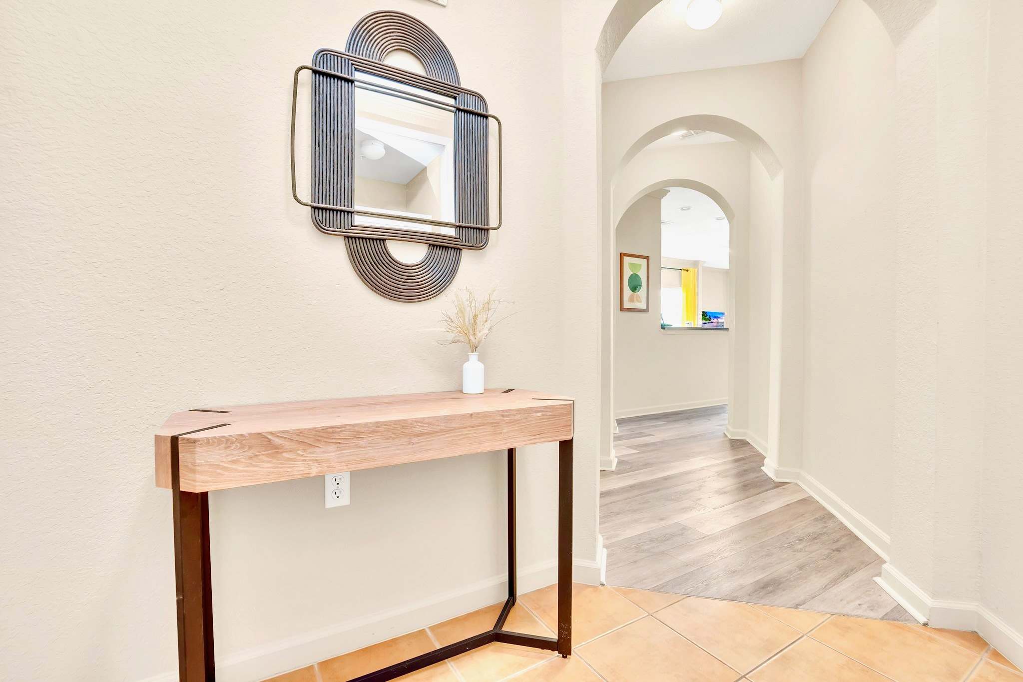 Inviting foyer with light-colored tile flooring, a modern wooden console table, and a decorative mirror, leading into a hallway with arched doorways.