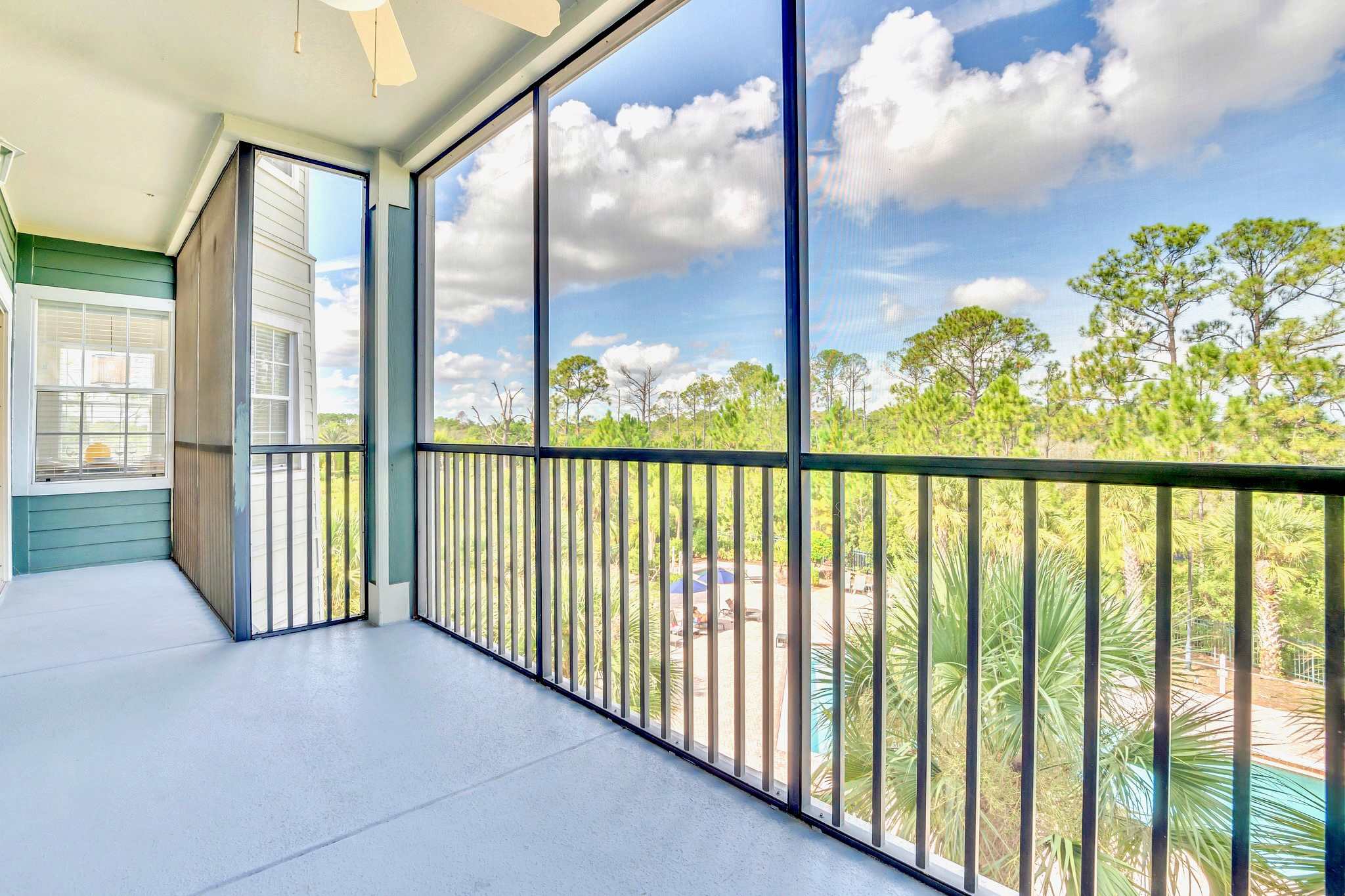 Spacious screened balcony offering expansive views of lush green trees and a distant pool area under a bright blue sky with white clouds.