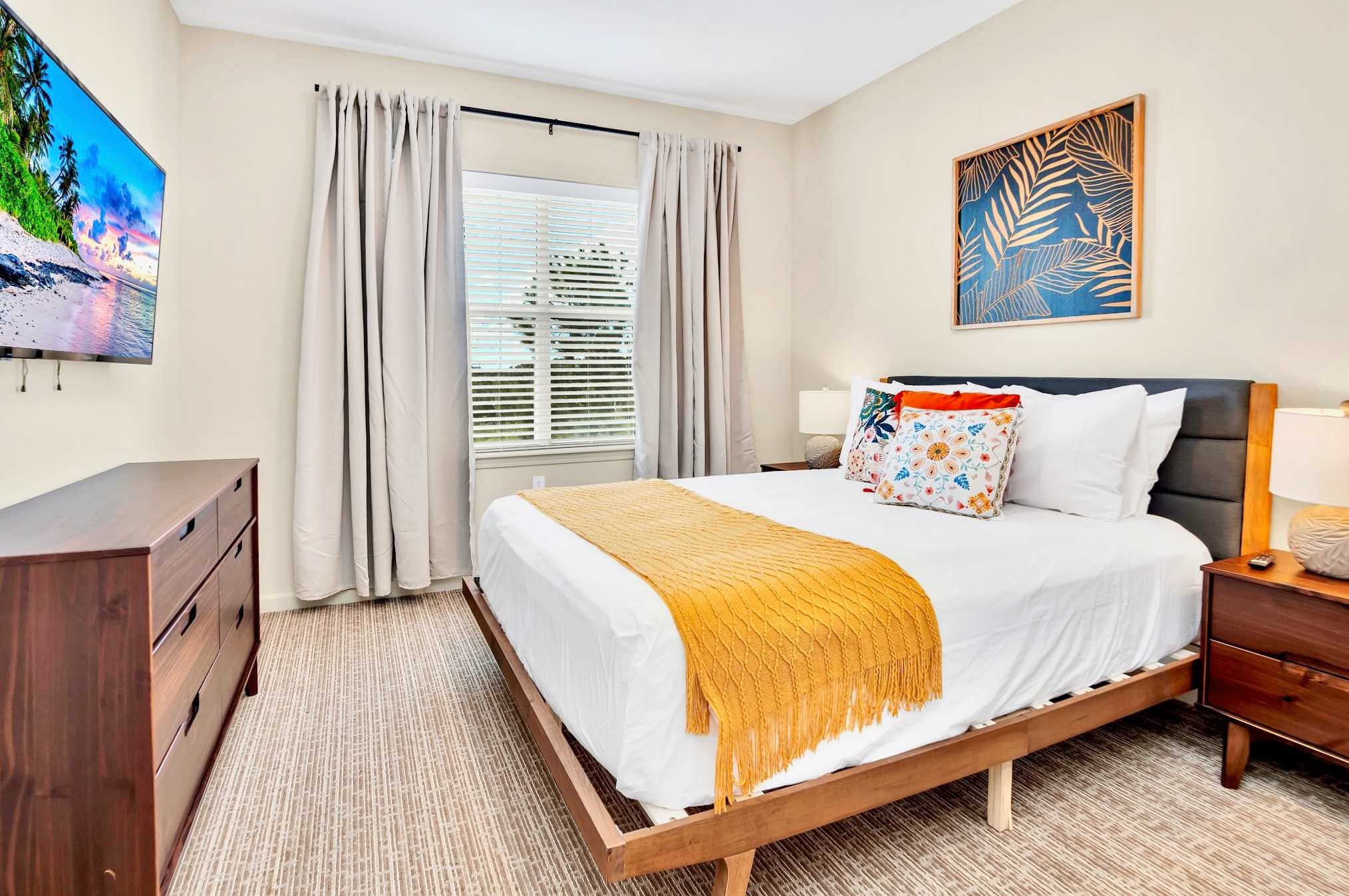 A cozy bedroom features a queen-sized bed with white linens and a yellow throw, flanked by a dark wood nightstand and a dresser with a wall-mounted TV. Light-colored curtains frame a window with blinds, and a large botanical print hangs above the bed.