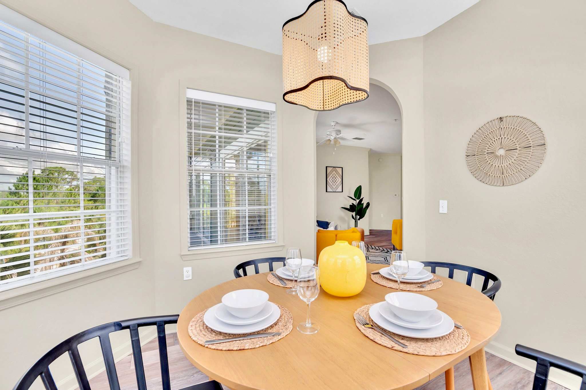 Cozy dining area featuring a round wooden table set for four with black chairs and a bright yellow vase centerpiece. A unique woven pendant light hangs above, complementing the natural light from two windows with blinds.