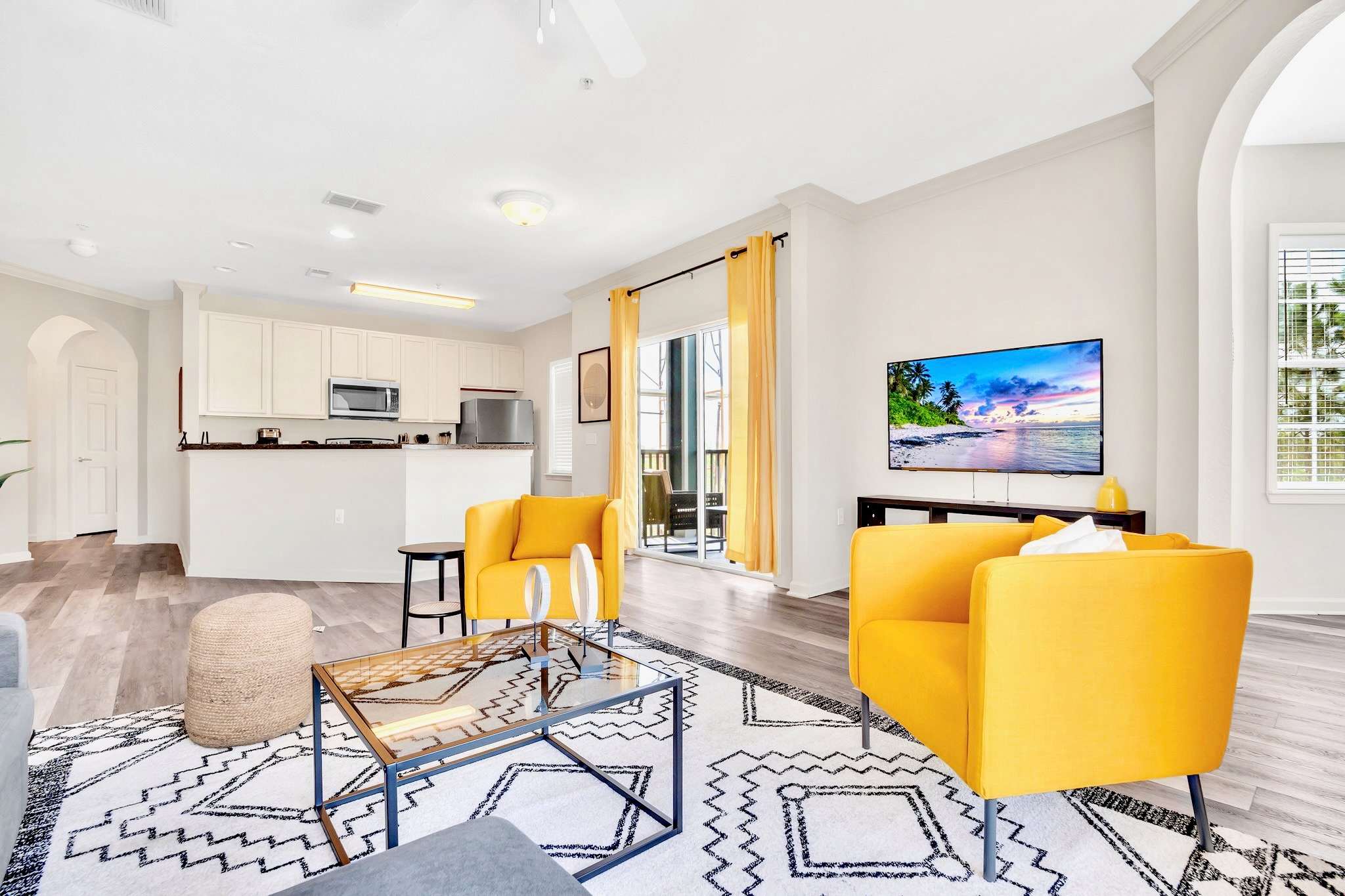 Spacious living area with a wall-mounted TV, two yellow armchairs, and a glass coffee table centered on a bold geometric rug. The kitchen's breakfast bar is visible in the background, connecting the spaces.