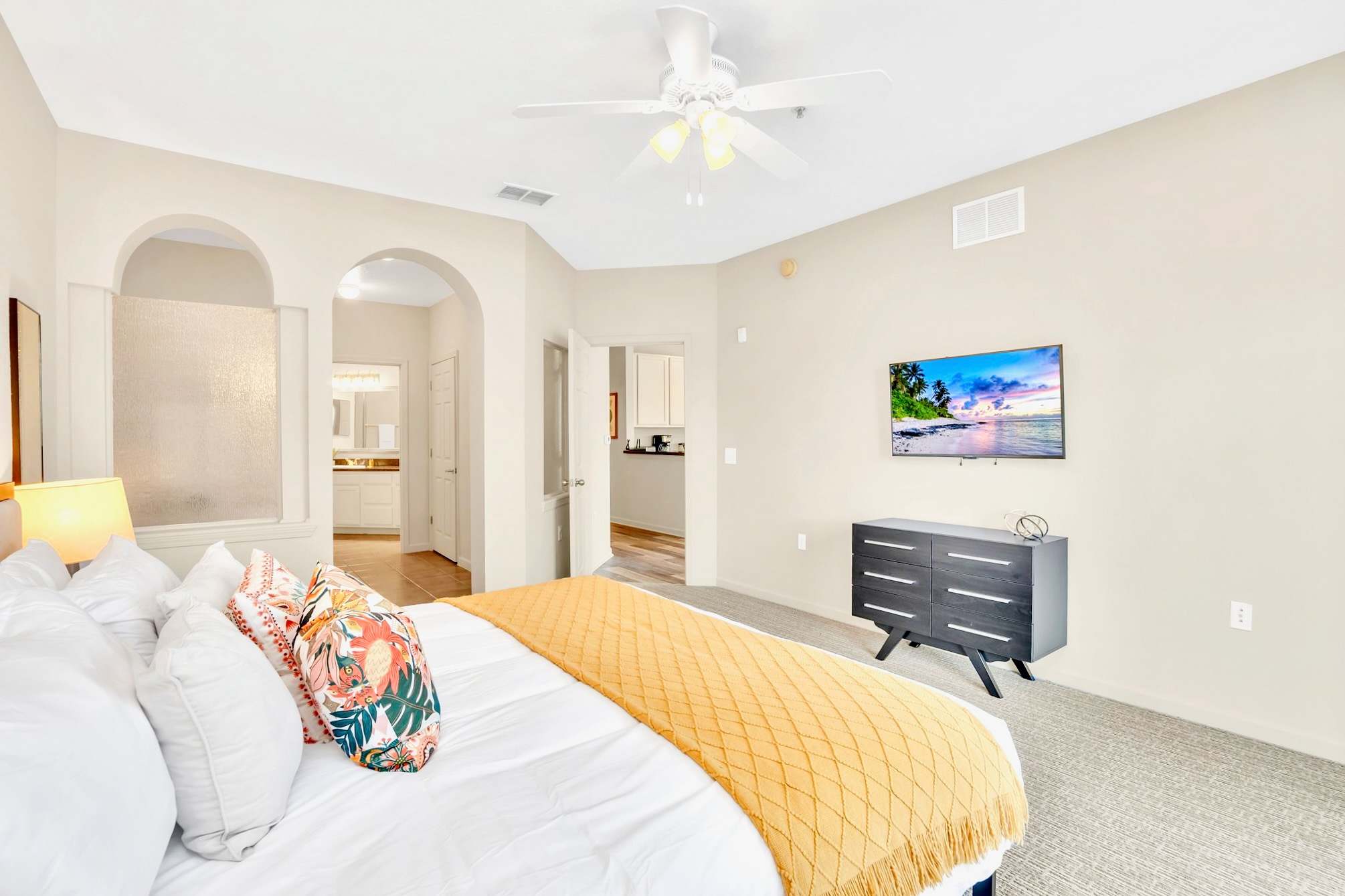 View of the bedroom showcasing the king-sized bed with colorful accent pillows and a wall-mounted TV opposite. A dark dresser sits below the TV, and an arched entryway leads to the en-suite bathroom.
