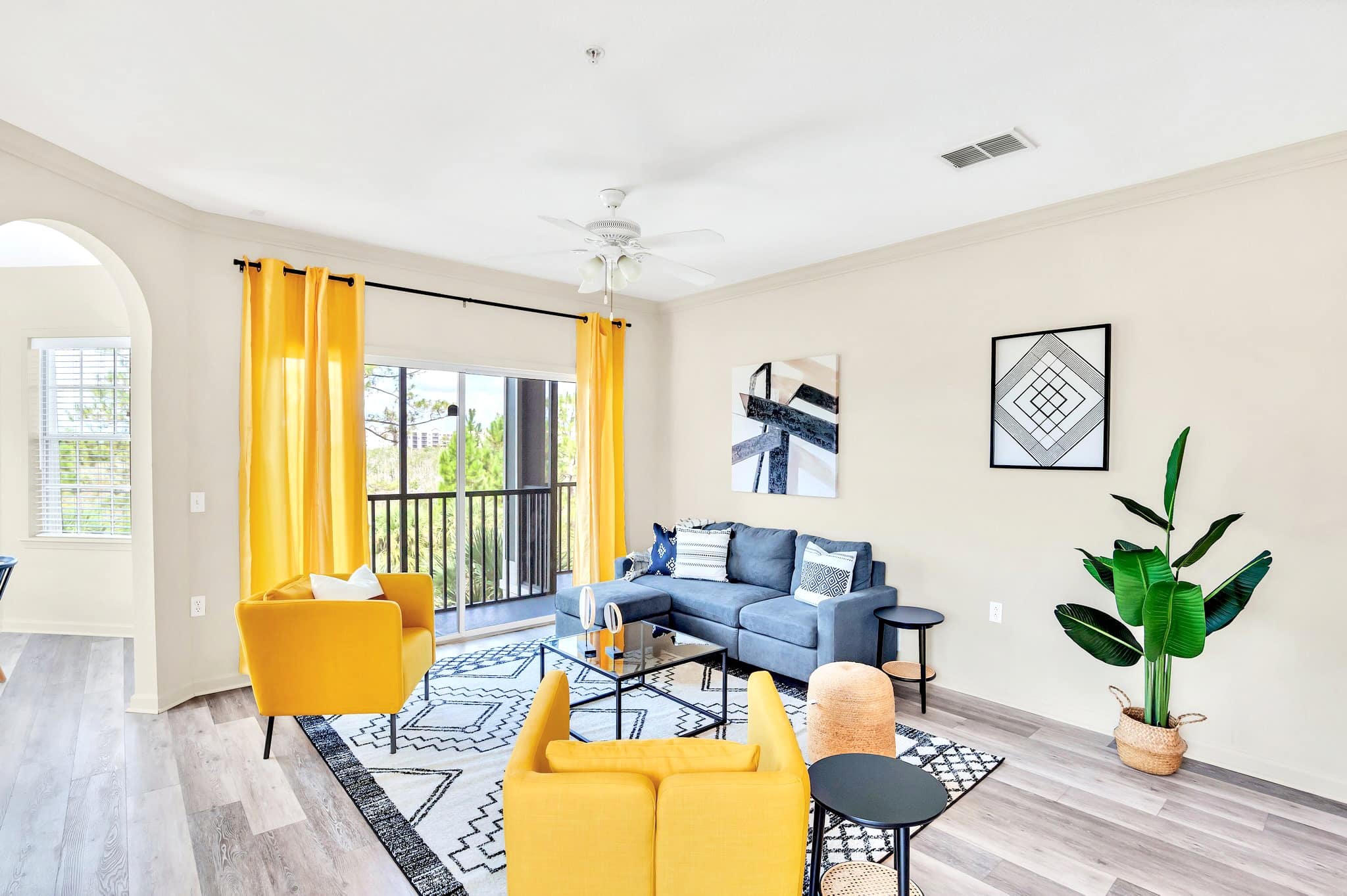 Bright living room featuring a grey sectional sofa, two vibrant yellow armchairs, and a glass-top coffee table on a black and white geometric rug. Yellow curtains frame a sliding glass door leading to a balcony.
