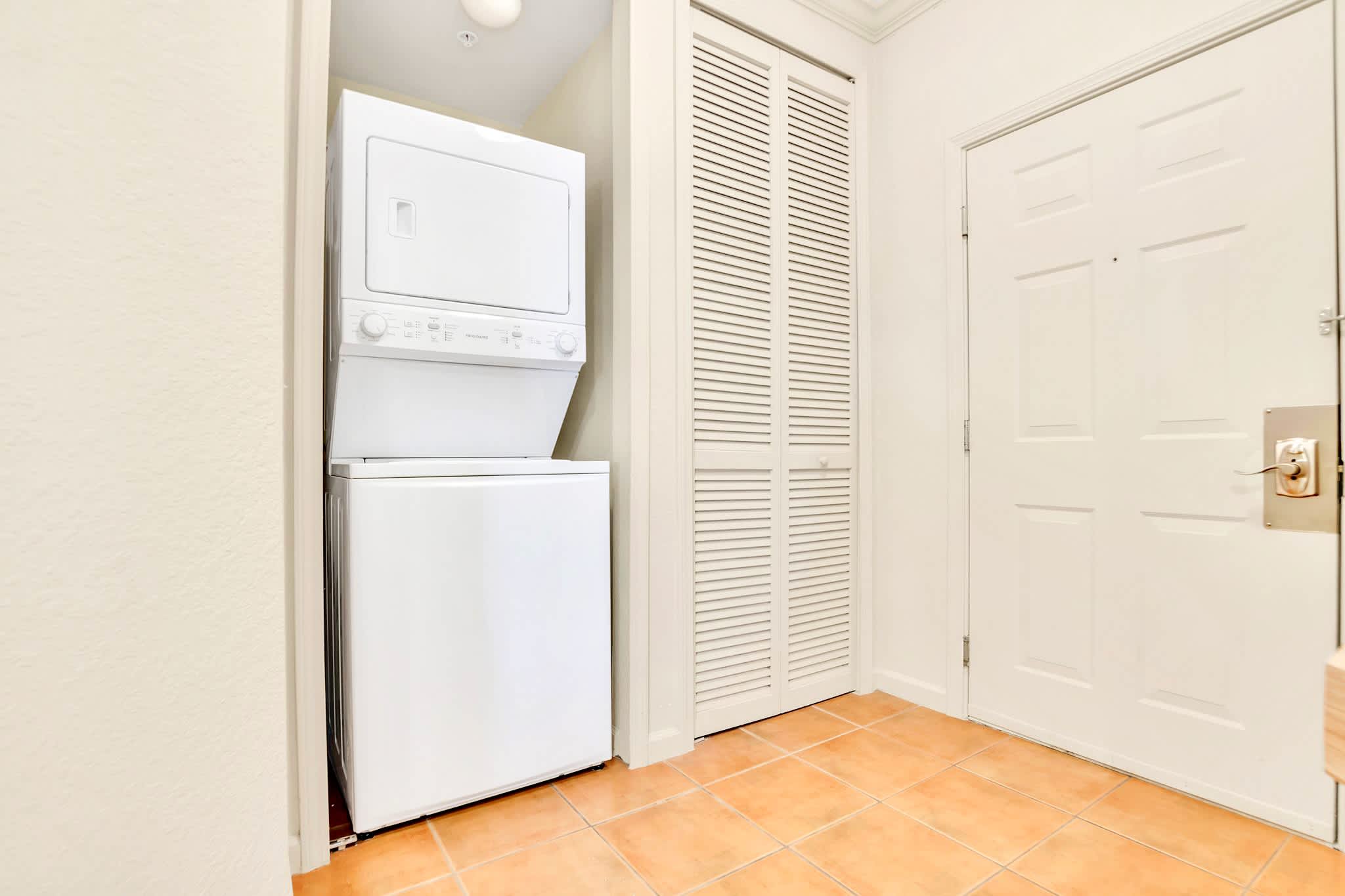 A compact laundry area features a stacked white washer and dryer unit, located next to a bi-fold closet door and a main entry door. The floor is tiled in a warm, earthy tone.